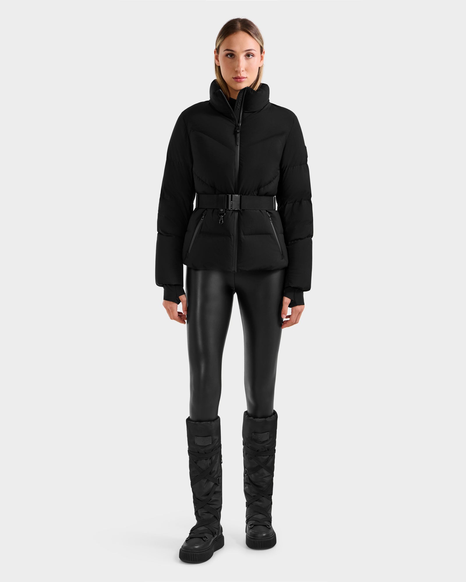 A woman faces forward against a light gray background, wearing Rudsak’s CHRISTELLE M - L125542 Black weather-resistant belted jacket, paired with black leggings and black quilted knee-high boots.