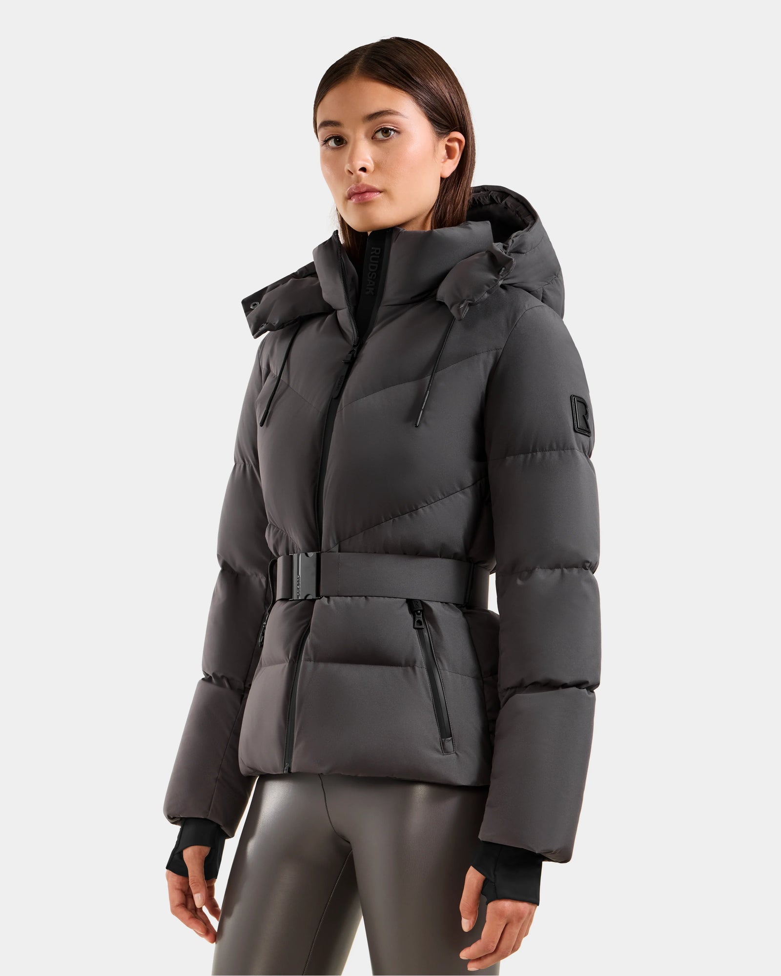 A woman wears the Rudsak CHRISTELLE M - L125542 Grey, a dark grey weather-resistant quilted down puffer jacket with hood and zippered pockets, paired with shiny dark leggings, against a plain light background.