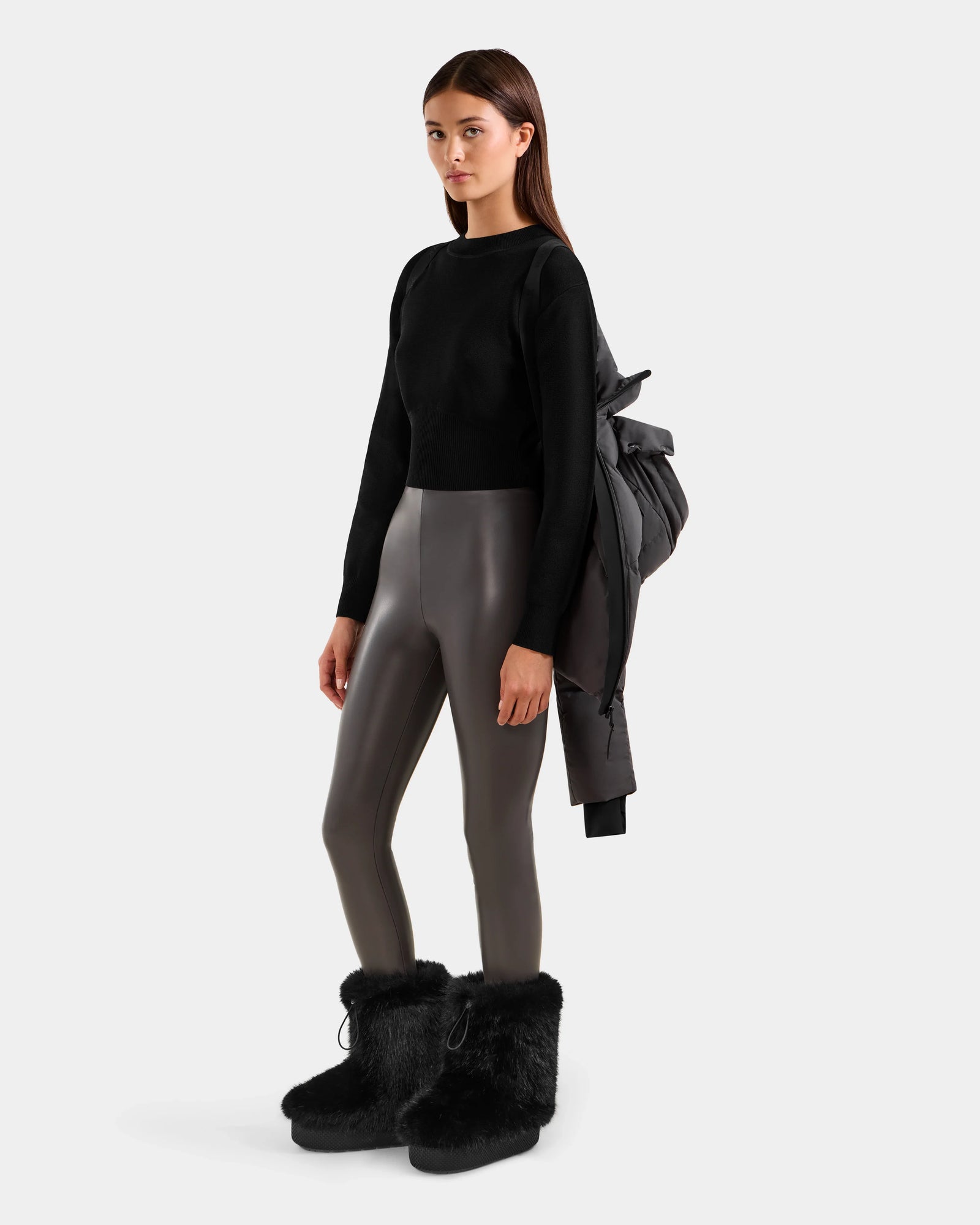 A woman poses against a plain background, wearing Rudsak's CHRISTELLE M - L125542 Grey quilted down puffer jacket draped over her left shoulder, paired with a black long-sleeve top, gray shiny leggings, and black furry boots.
