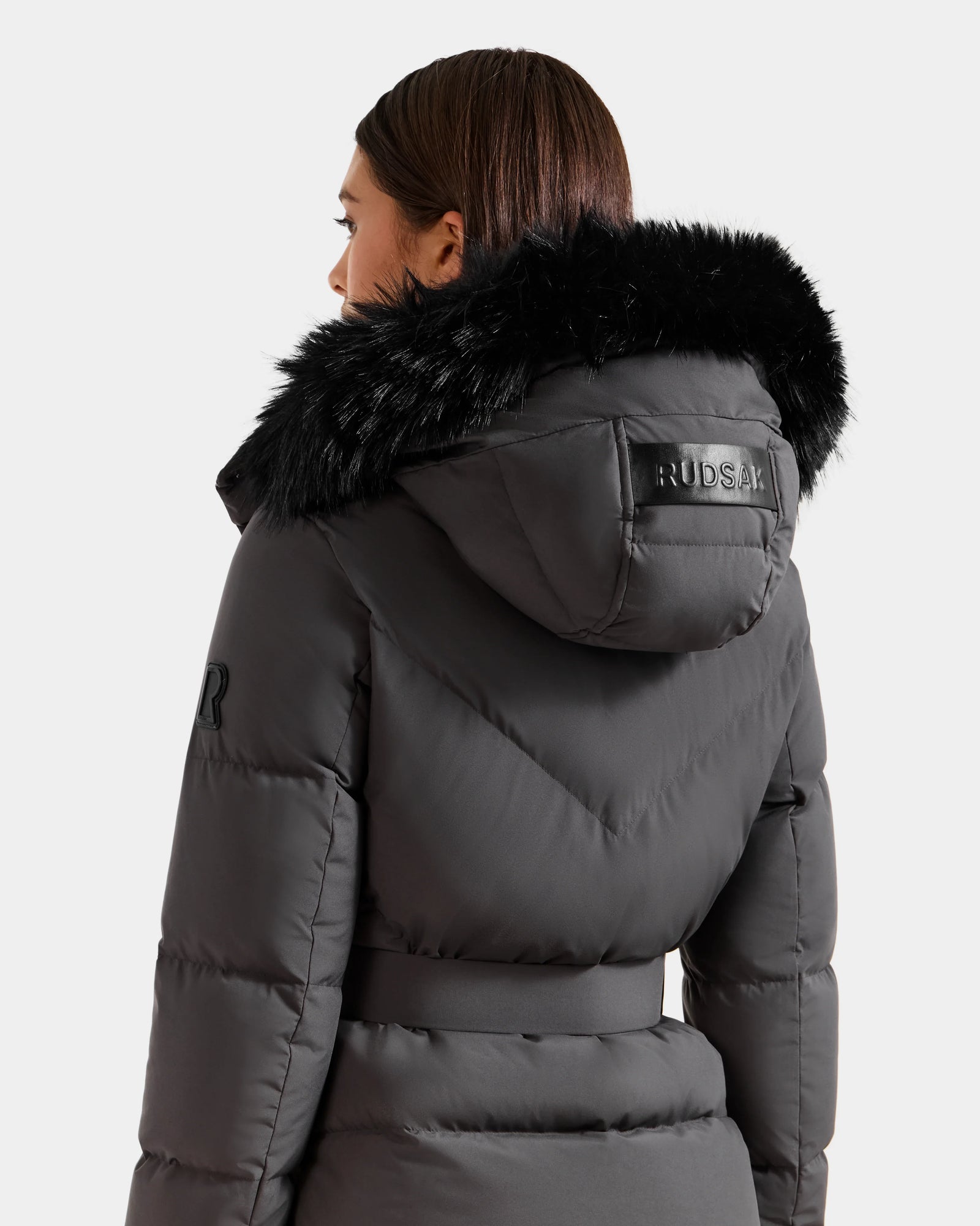 A woman with straight brown hair wears the Rudsak CHRISTELLE M - L125542 Grey, a weather-resistant quilted down puffer with a black faux fur-trimmed hood. "RUDSAK" is on the hood's back and a logo appears on the upper arm.