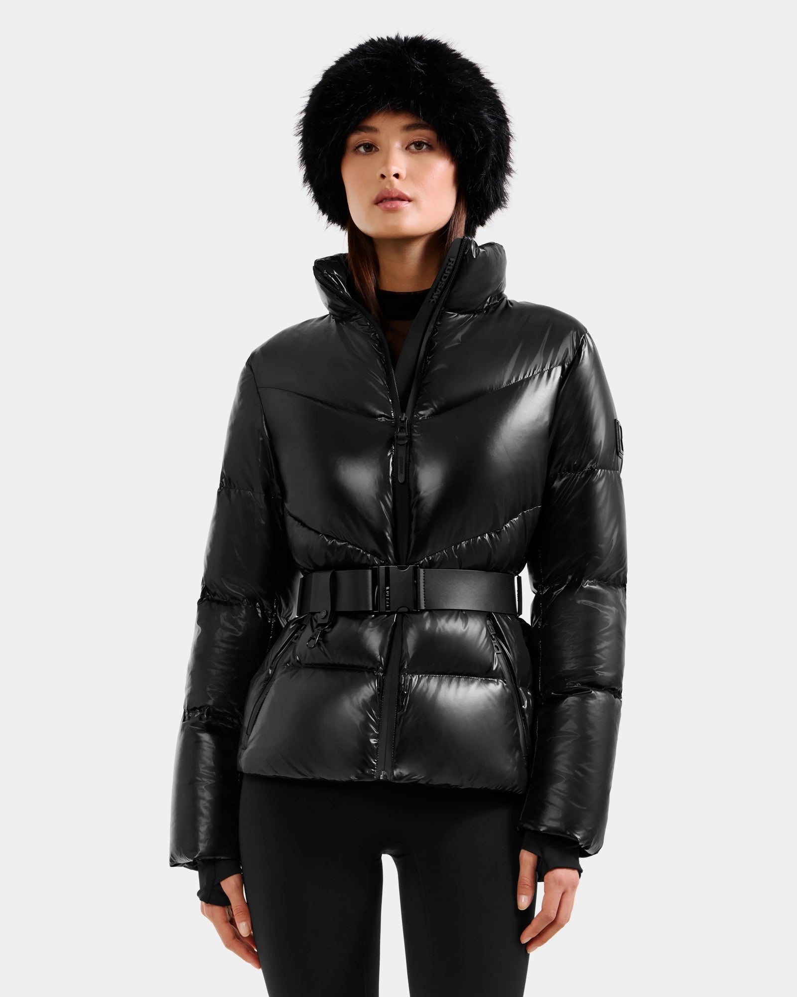 A woman with straight brown hair wears the Rudsak CHRISTELLE S - L125544 Black women’s down puffer jacket with a belt, paired with a faux fur trim hat, standing against a plain light background and looking slightly upward.