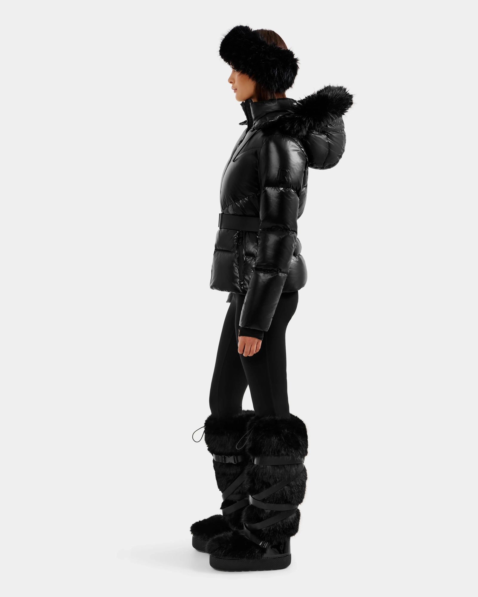 A woman in the Rudsak CHRISTELLE S - L125544 Black women’s down puffer with faux fur trim, paired with a black fur headband, black leggings, and tall black fur boots, stands in profile against a plain background.