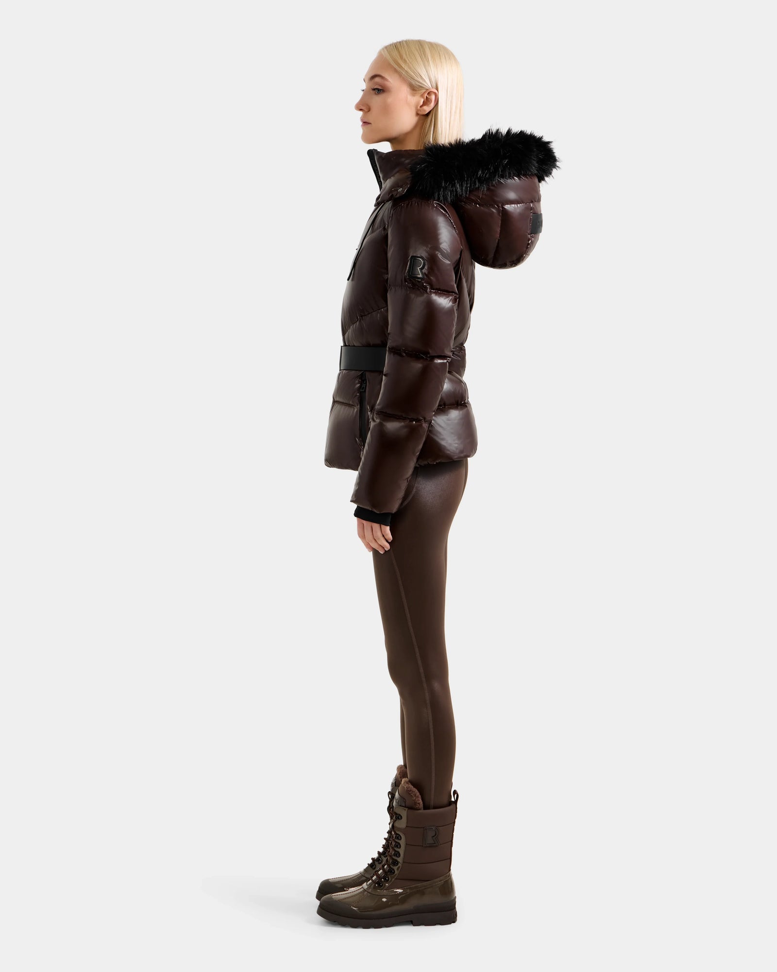 A woman with straight blonde hair stands in profile wearing the Rudsak CHRISTELLE S - L125544 Espresso women's shiny dark brown down puffer jacket with a faux fur trim hood, against a plain light background.