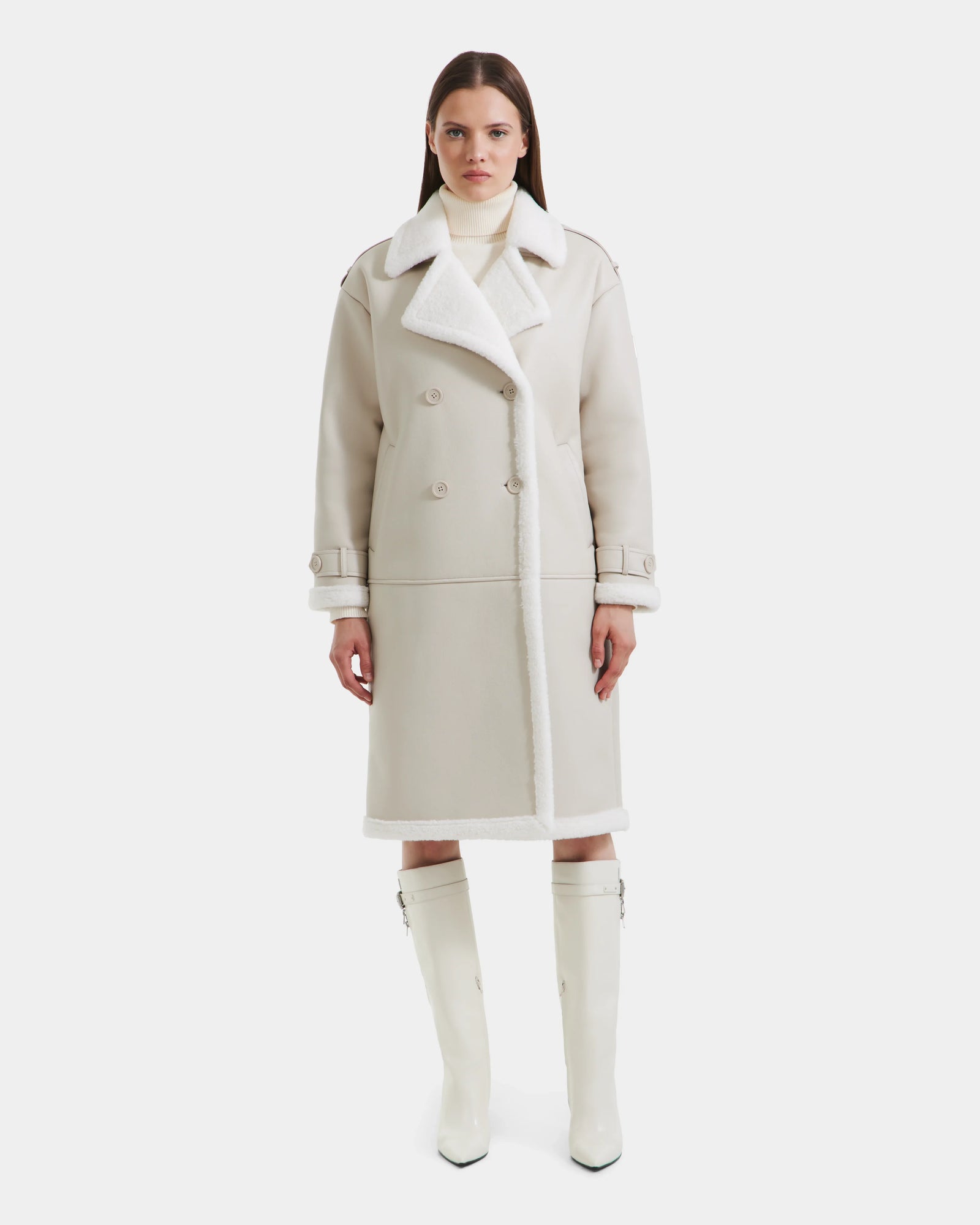 A woman faces forward in the Rudsak DARCY - L125539 Vanilla long, light beige winter coat with wide lapels and double-breasted buttons, paired with a white turtleneck and knee-high white boots against a plain white background.