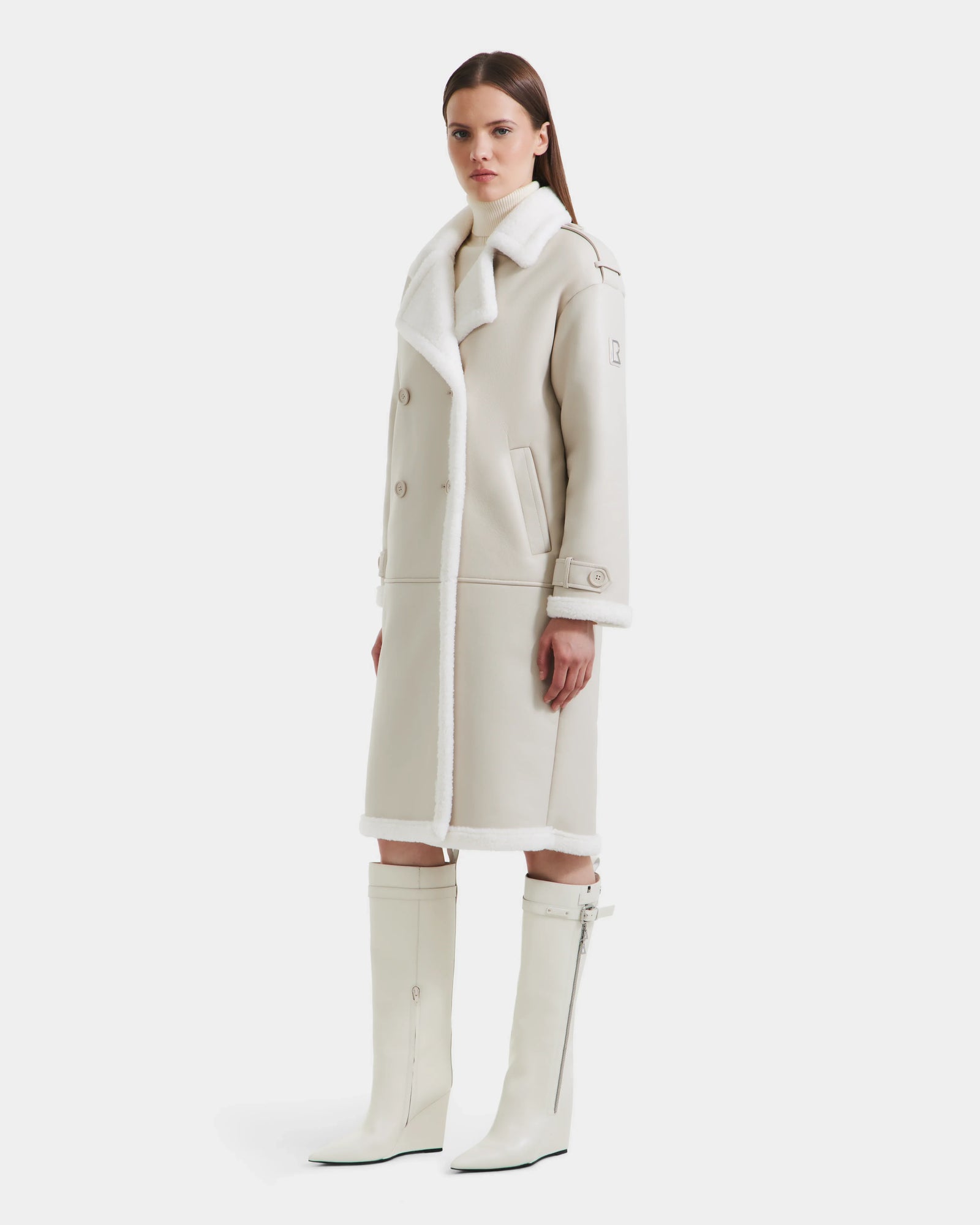A woman wears the Rudsak DARCY - L125539 Vanilla long winter coat with white fur trim and matching knee-high boots, facing forward with a neutral expression against a plain white background.