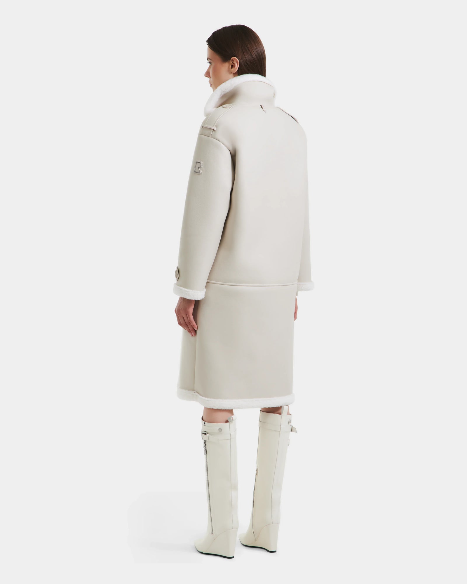 A woman stands facing away in the DARCY - L125539 Vanilla winter coat by Rudsak, its elegant faux sherpa teddy texture and high collar highlighted against a white background, paired with matching knee-high boots.