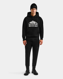 A person stands against a plain background wearing Rudsak’s DARWIN - M525502 Black cotton-blend fleece men’s hoodie with Ski Culture and mountain graphic, paired with a black beanie, black pants, and black boots.