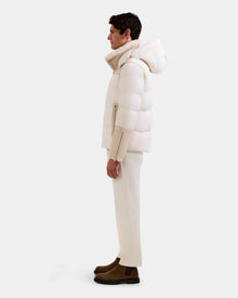 A man stands in profile against a plain background, wearing the Rudsak DAVID - M125508 Vanilla men's shearling puffer jacket with a hood, matching off-white pants, and brown suede boots.