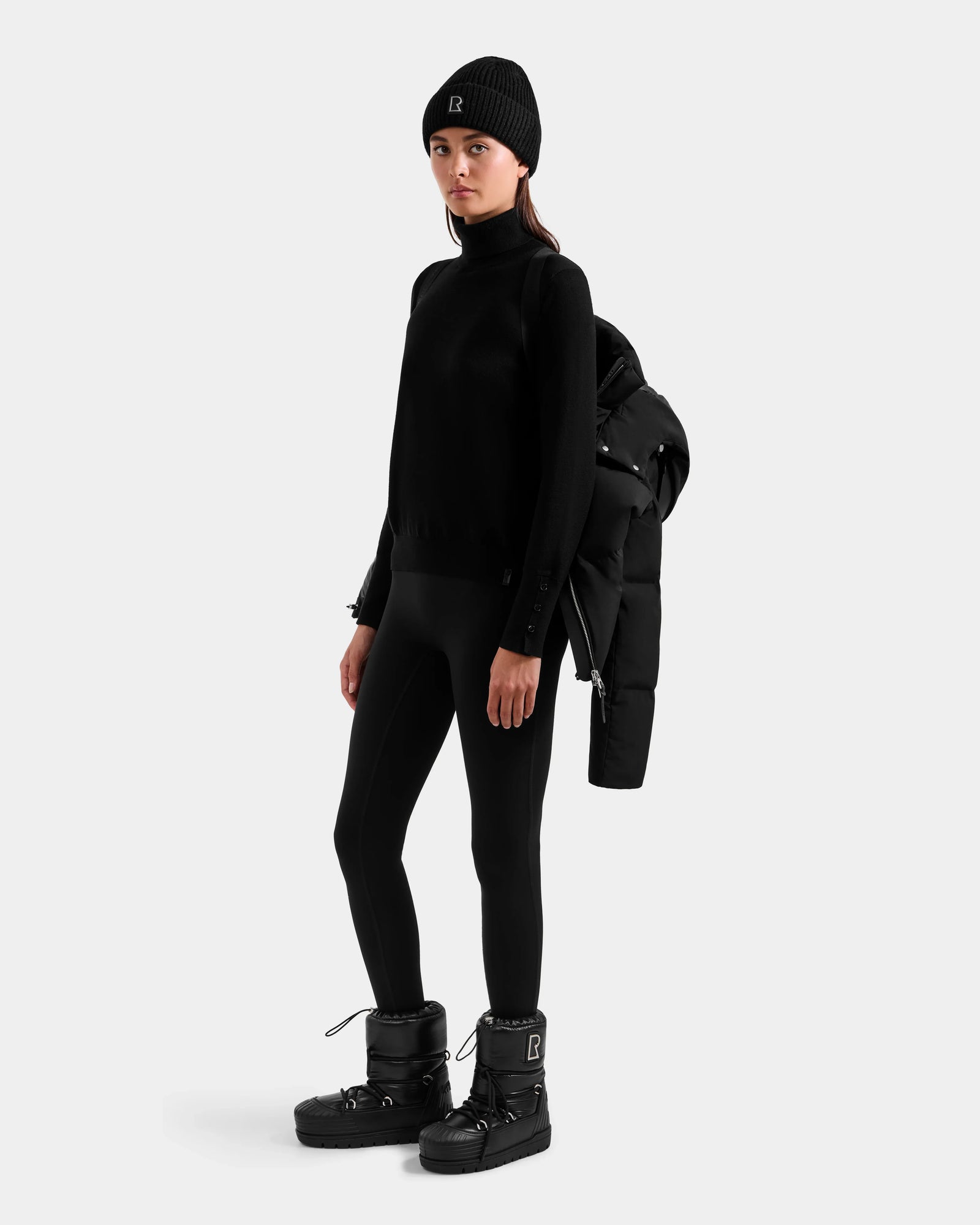 A woman in a black beanie, sweater, and leggings poses in Rudsak boots with the DELILAH M - L125538 Black women's down puffer draped over her shoulder against a plain light gray background.