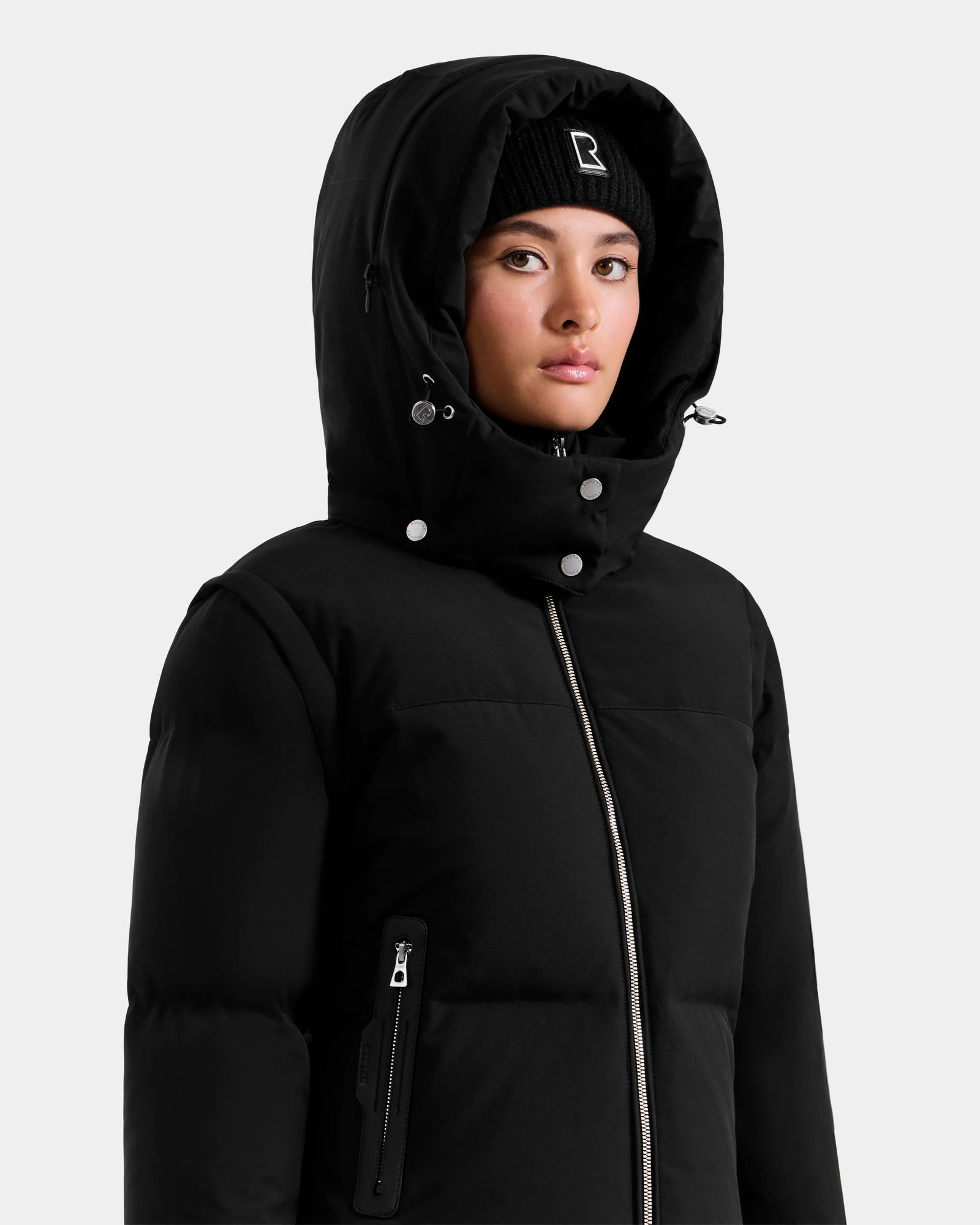 A person wearing the Rudsak DELILAH M - L125538 Black women’s down puffer with the hood up and a black beanie, looking slightly to the side against a plain light background.