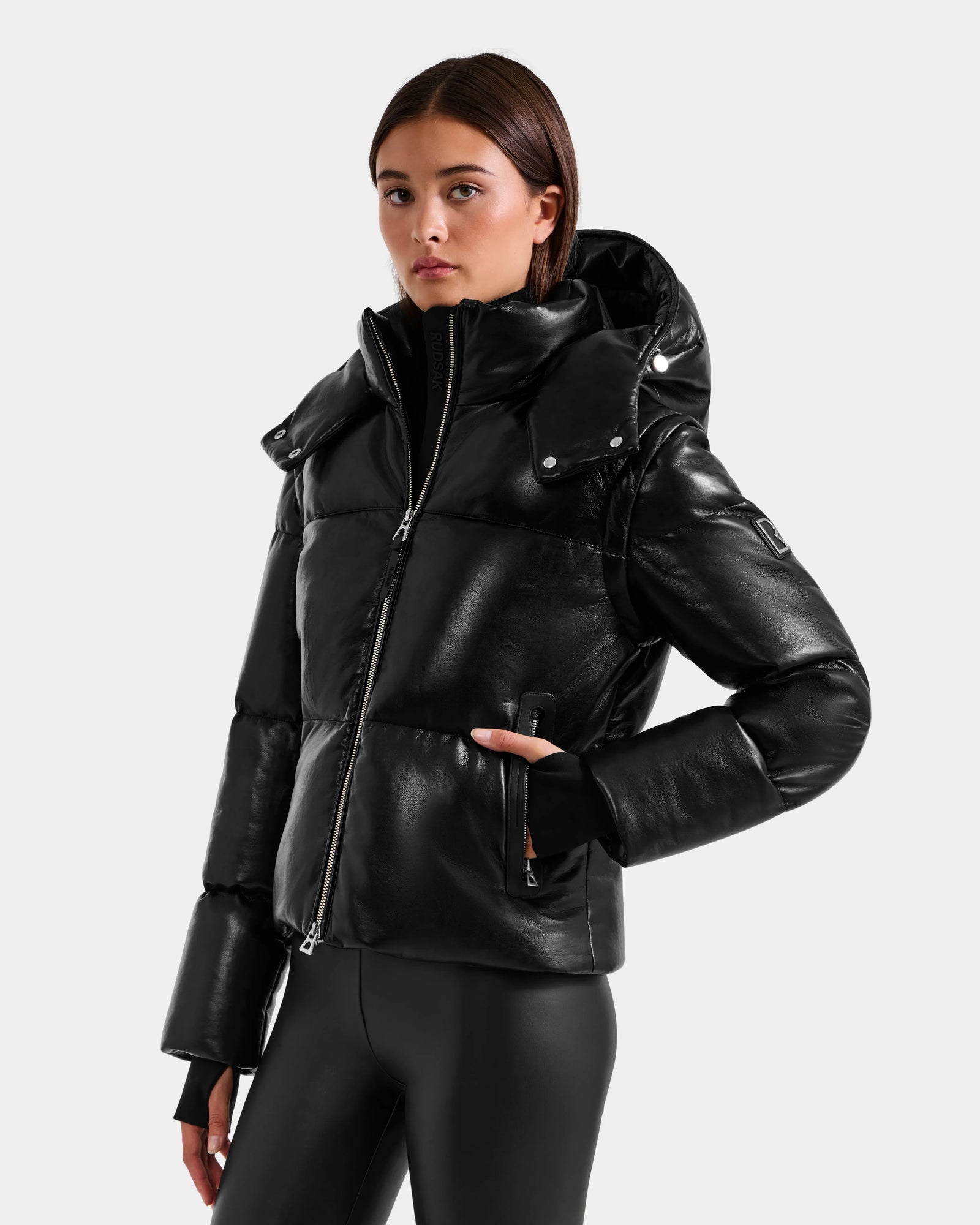 A woman wearing the Rudsak DELILAH PX - L125537 Black vegan-leather puffer jacket with a hood and black leggings stands against a plain light gray background, confidently looking at the camera with one hand in her pocket.