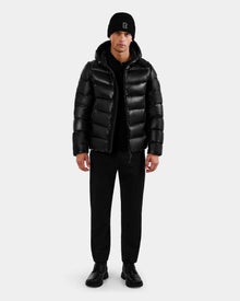 A man faces forward wearing the Rudsak DEMIAN S-R - M123505 Black down puffer jacket, paired with black pants, shoes, and a beanie with a small logo. The background is plain and light-colored.
