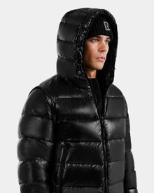 A person wears the Rudsak DEMIAN S-R - M123505 Black men’s down puffer jacket with hood up and a black beanie, looking sideways against a plain light background. This stylish coat uses RDS-certified down for warmth and ethical comfort.