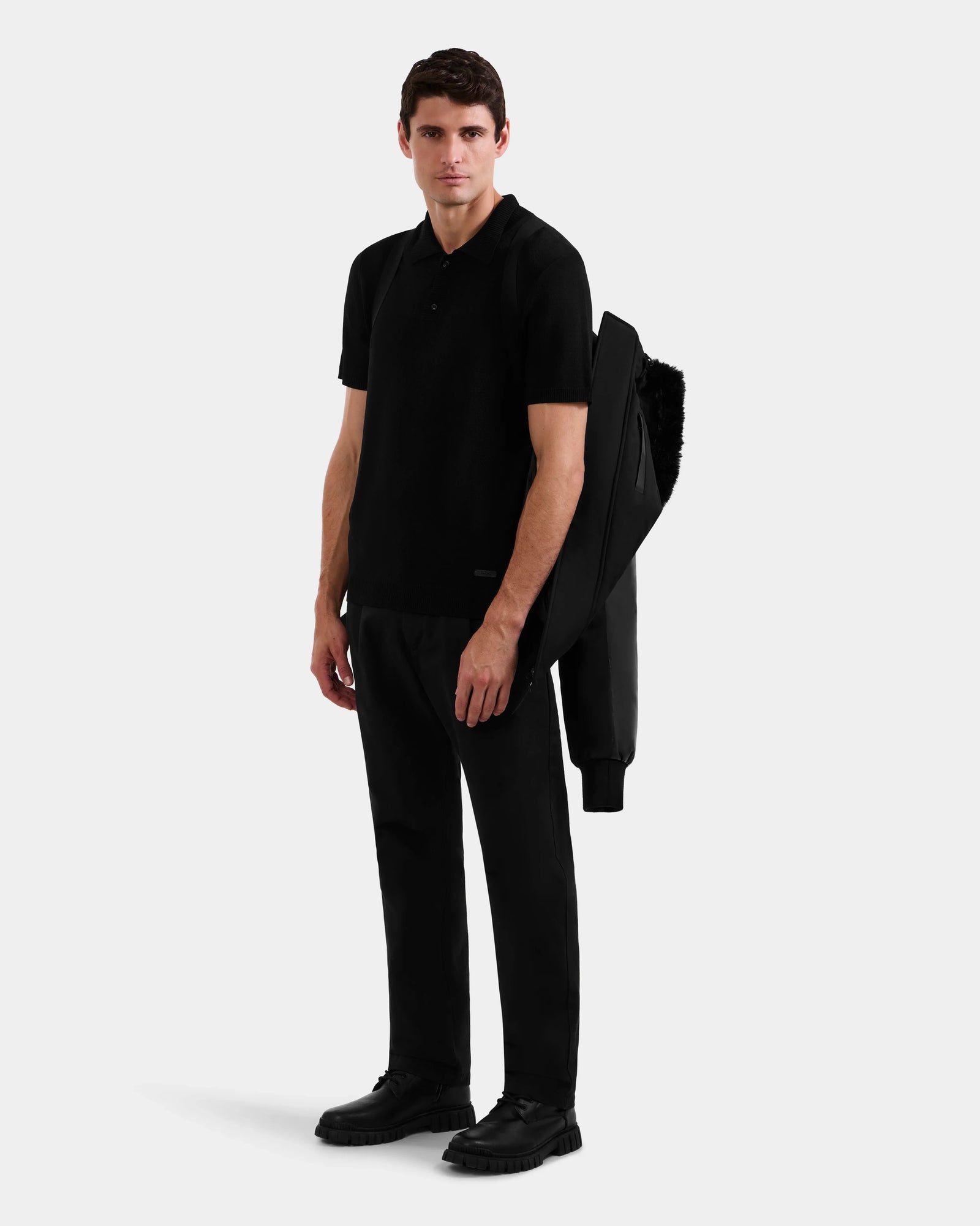 A man stands against a plain background, wearing all black and holding the Rudsak DERON - M125539 Black men’s down bomber with vegan-leather sleeves over his left arm, looking straight ahead with a neutral expression.