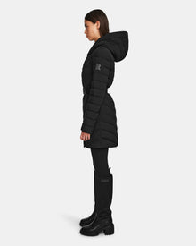 A woman stands in profile wearing the Rudsak DITA - L124556 Black quilted down puffer coat with a hood, paired with black leggings and tall black boots against a plain white background.