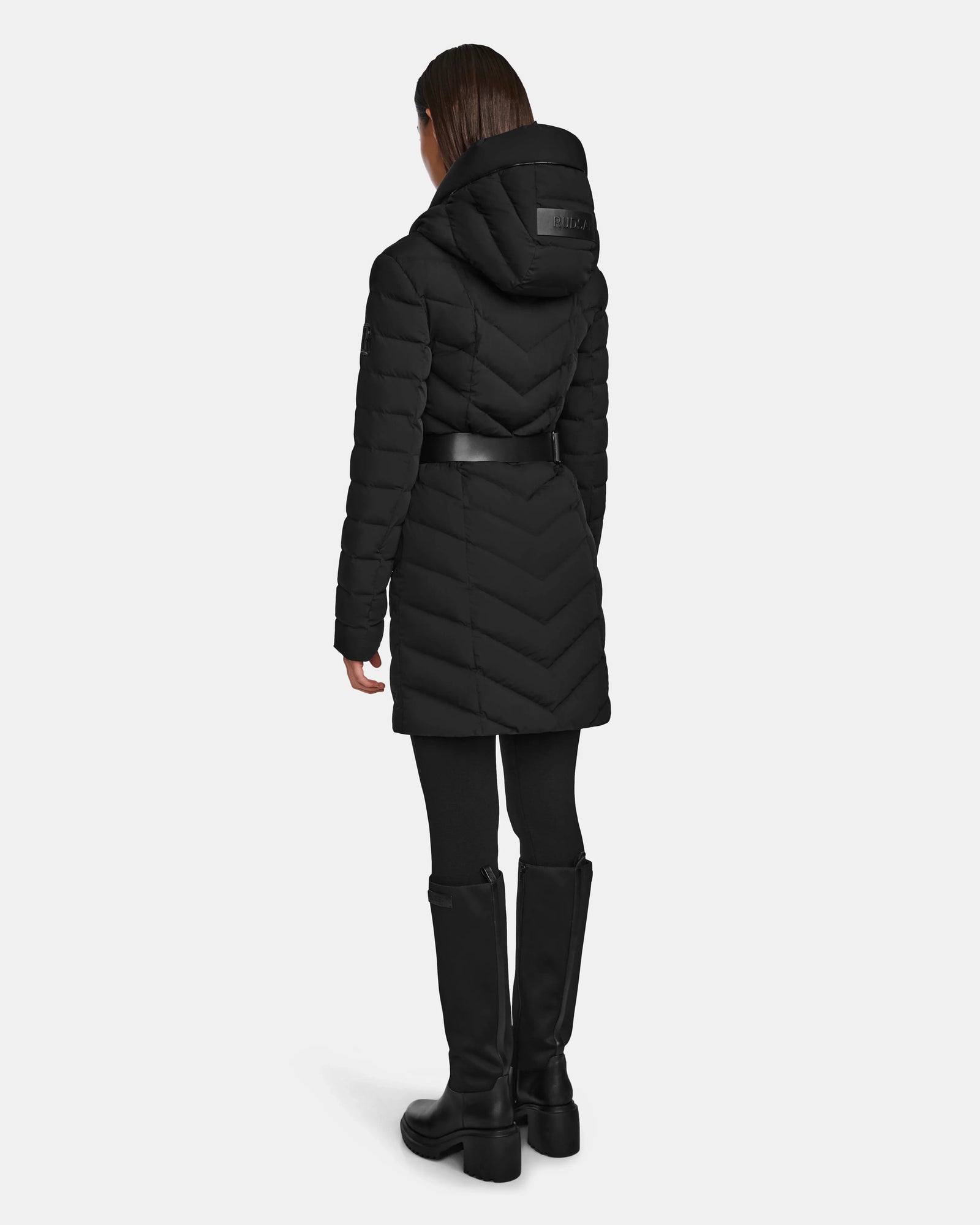 A person wearing the Rudsak DITA - L124556 Black women's quilted down puffer with a belt, black leggings, and black knee-high boots stands with their back to the camera against a plain light background.