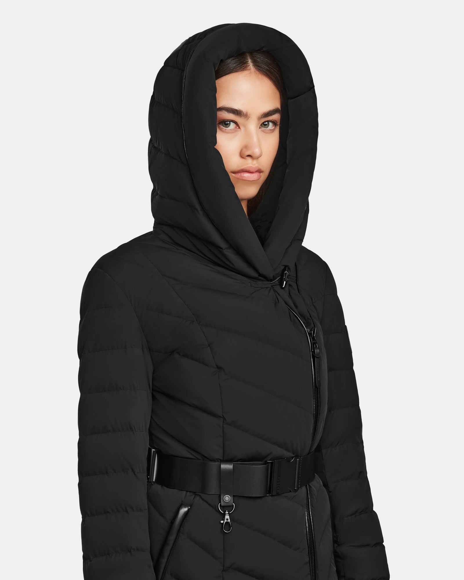 A woman in the Rudsak DITA - L124556 Black knee-length puffer jacket with a black belt and large hood stands against a light background, gazing slightly to the side with a neutral expression.