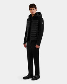 A man stands against a plain light gray background, wearing the Rudsak DURIN M - M125530 Black men's hooded jacket with waterproof zippers and lightweight down insulation, paired with black pants and shoes. He faces slightly to the side, expression neutral.