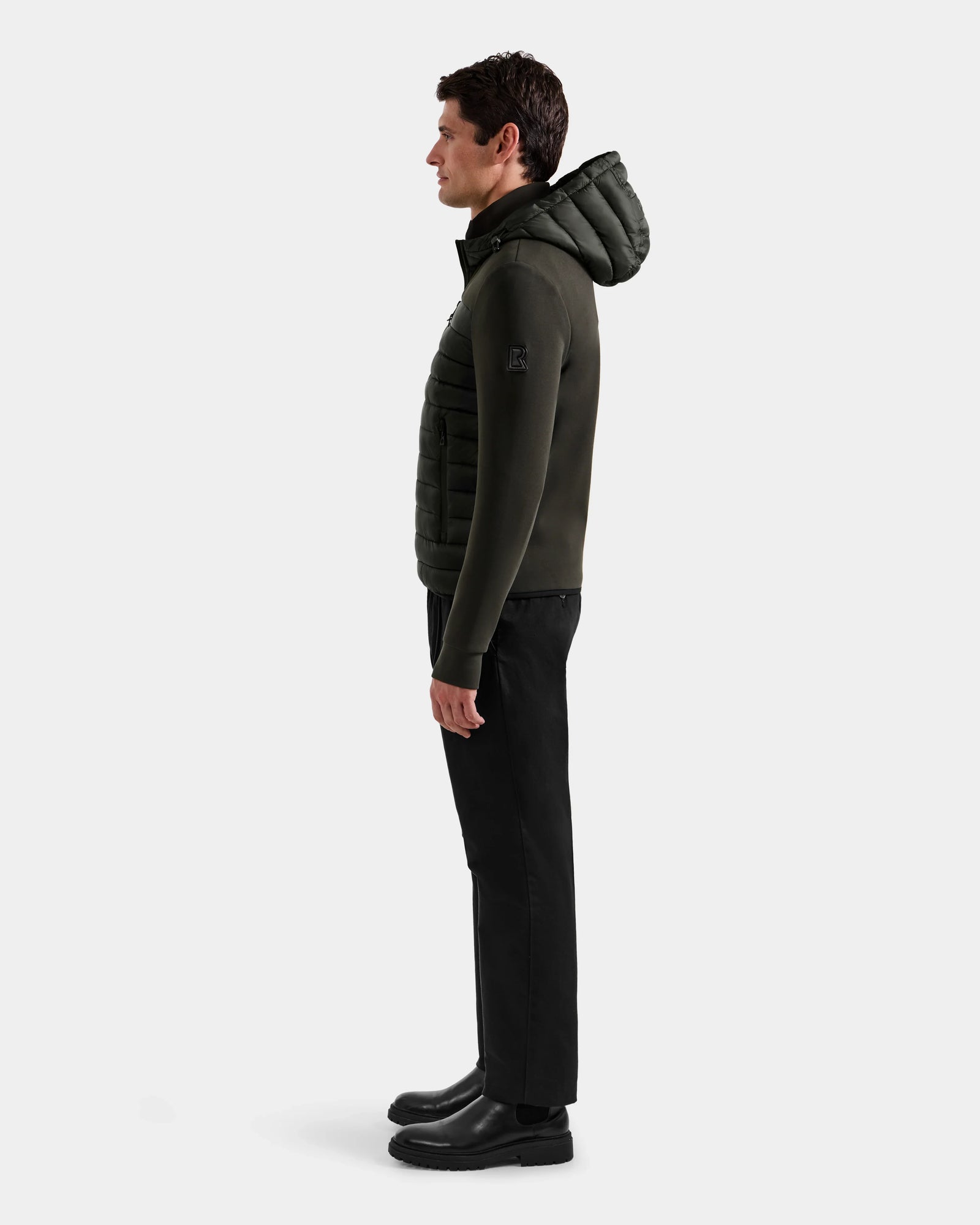 A man stands in profile against a plain background, wearing the Rudsak DURIN M - M125530 dark olive men’s hooded jacket with lightweight down insulation, paired with black pants and boots. He faces left with his arms relaxed at his sides.