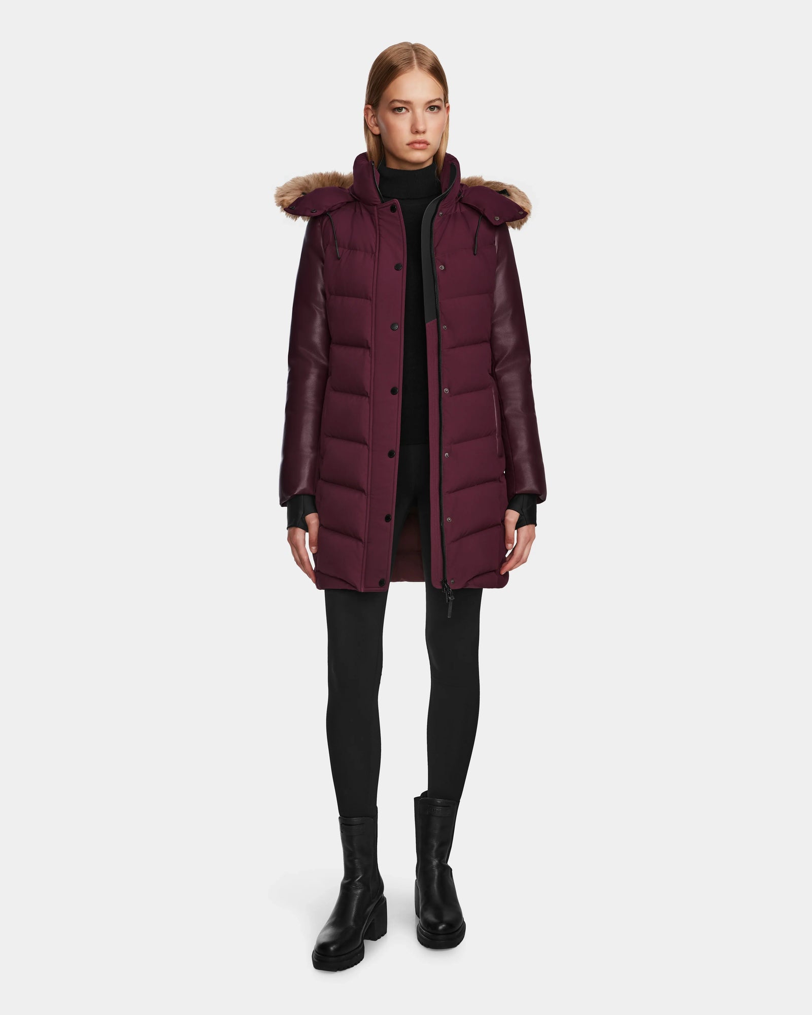 A woman wears the Rudsak ELSA - L123535 Wine quilted down puffer with a fur-trimmed hood, styled with black leggings, a turtleneck, and ankle boots against a plain white background.