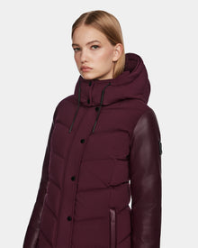 A woman with straight blonde hair wears the Rudsak ELSA - L123535 Wine women’s quilted down puffer jacket with leather sleeves and a hood, standing against a plain light background.