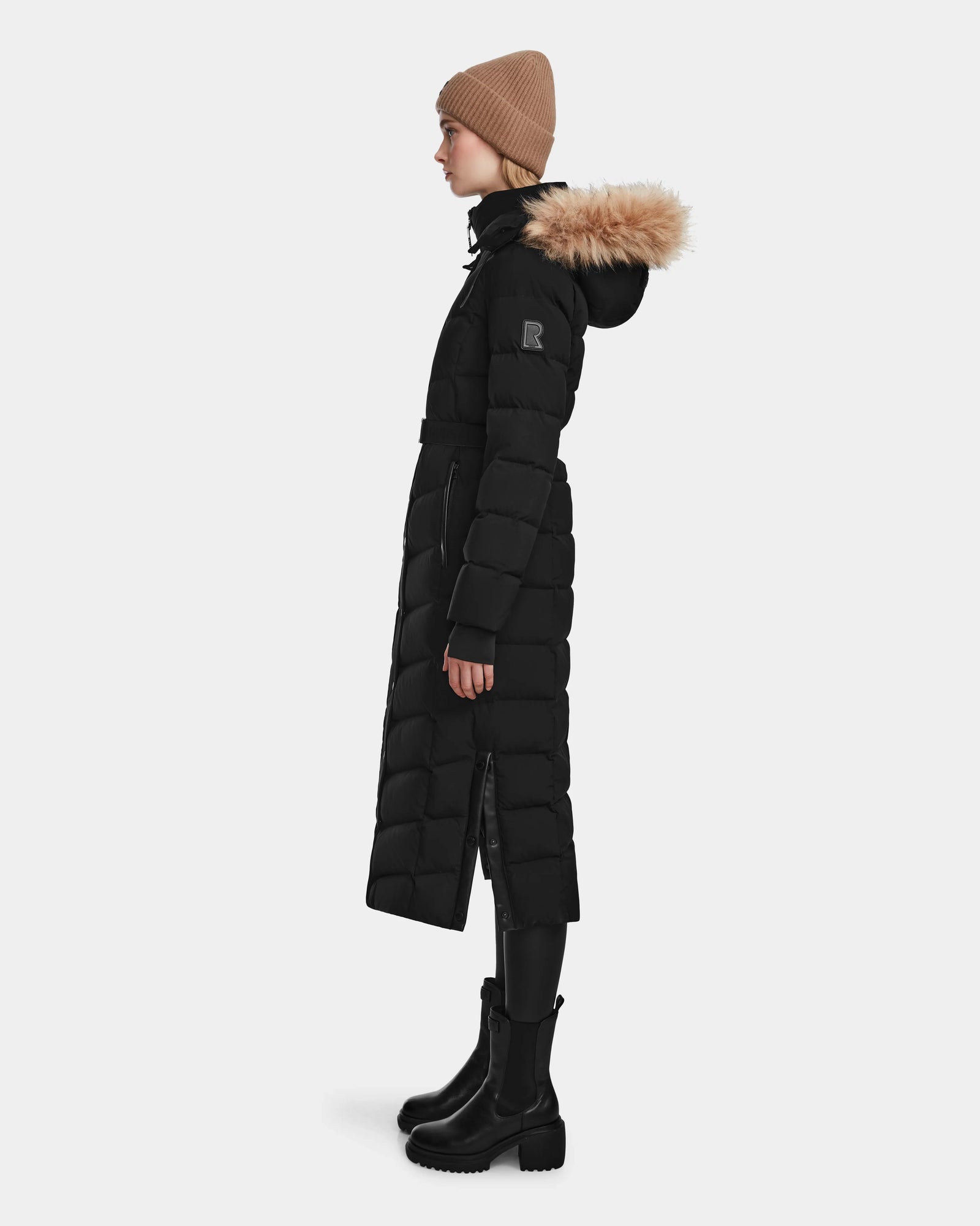 A person in profile wearing the Rudsak ELSIE M - L123517 Black/Natural women’s maxi down puffer with a fur-trimmed hood, black leggings, chunky boots, and a tan knit beanie, standing against a plain white background.