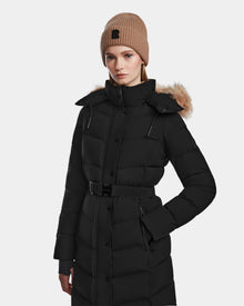 A woman models the Rudsak ELSIE M - L123517 Black/Natural maxi down puffer with belt and fur-lined hood, wearing a tan knit beanie and posing against a plain light background.