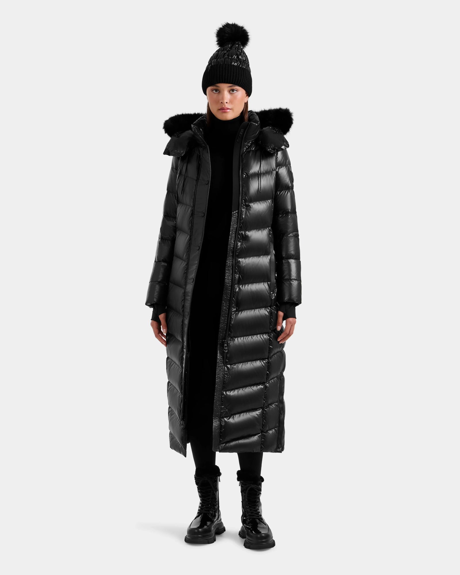 A person wears the Rudsak ELSIE S - L124536 Black women’s maxi down puffer with water-resistant fabric and fur-trimmed hood, paired with a black pom-pom beanie, black pants, and boots against a plain light background.