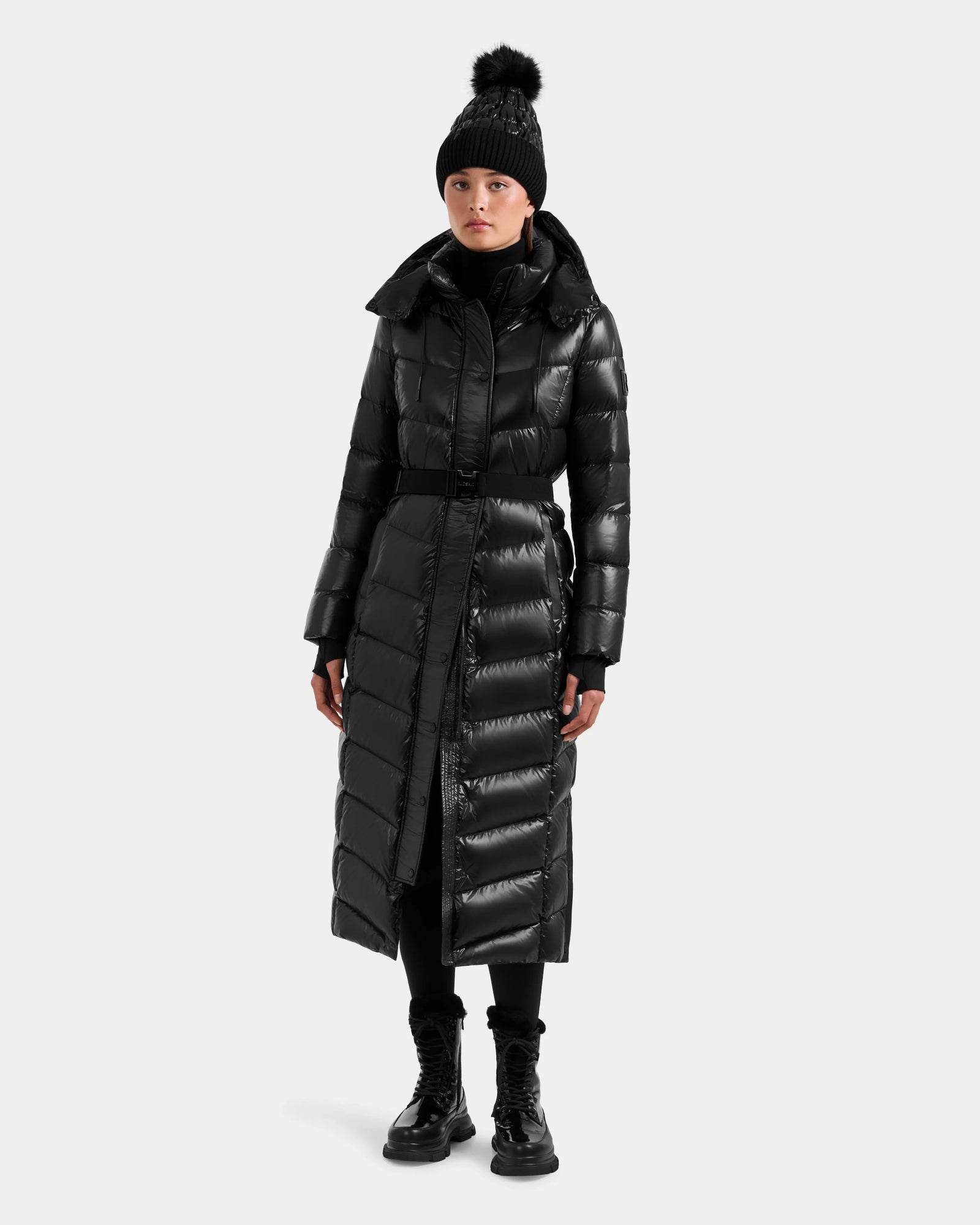 A person stands against a plain light background wearing the Rudsak ELSIE S - L124536 Black women’s maxi down puffer with water-resistant, RDS-certified down, paired with a black pom-pom knit hat, black pants, and lace-up boots.