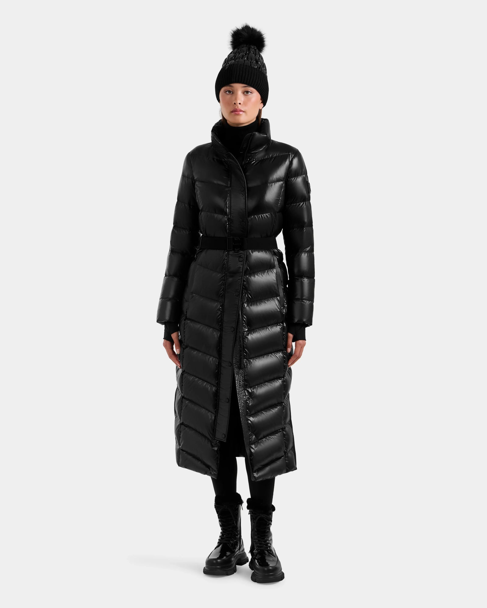A person wears the Rudsak ELSIE S - L124536 Black women’s maxi down puffer, made with water-resistant fabric and RDS-certified down, styled with a black beanie, turtleneck, pants, and lace-up boots against a plain light background.