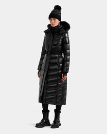 A person wearing the Rudsak ELSIE S - L124536 Black women’s maxi down puffer, paired with a black pom-pom beanie, boots, and leggings, stands against a plain light background.