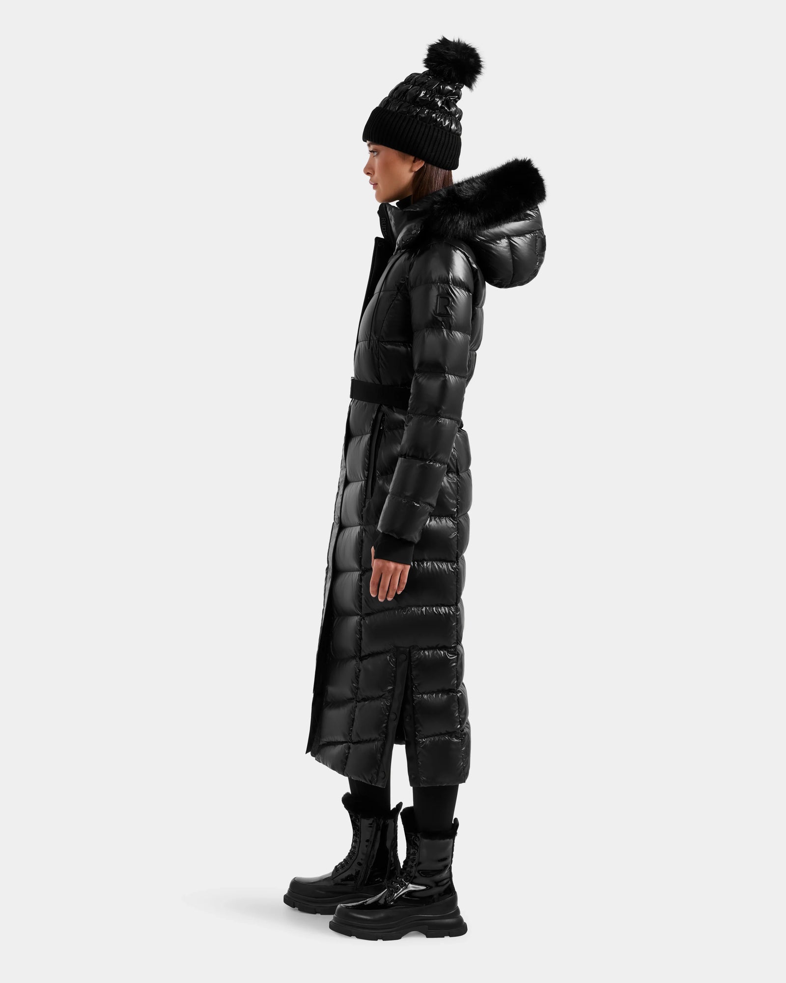 A woman in profile wears the Rudsak ELSIE S - L124536 Black maxi down puffer with a fur-lined hood, black winter boots, and a pom-pom beanie against a plain white background. The coat is insulated with RDS-certified down for warmth.