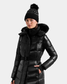 A woman in the Rudsak ELSIE S - L124536 Black water-resistant puffer coat with a fur-lined hood and a black knit pom-pom beanie, standing against a plain light background.
