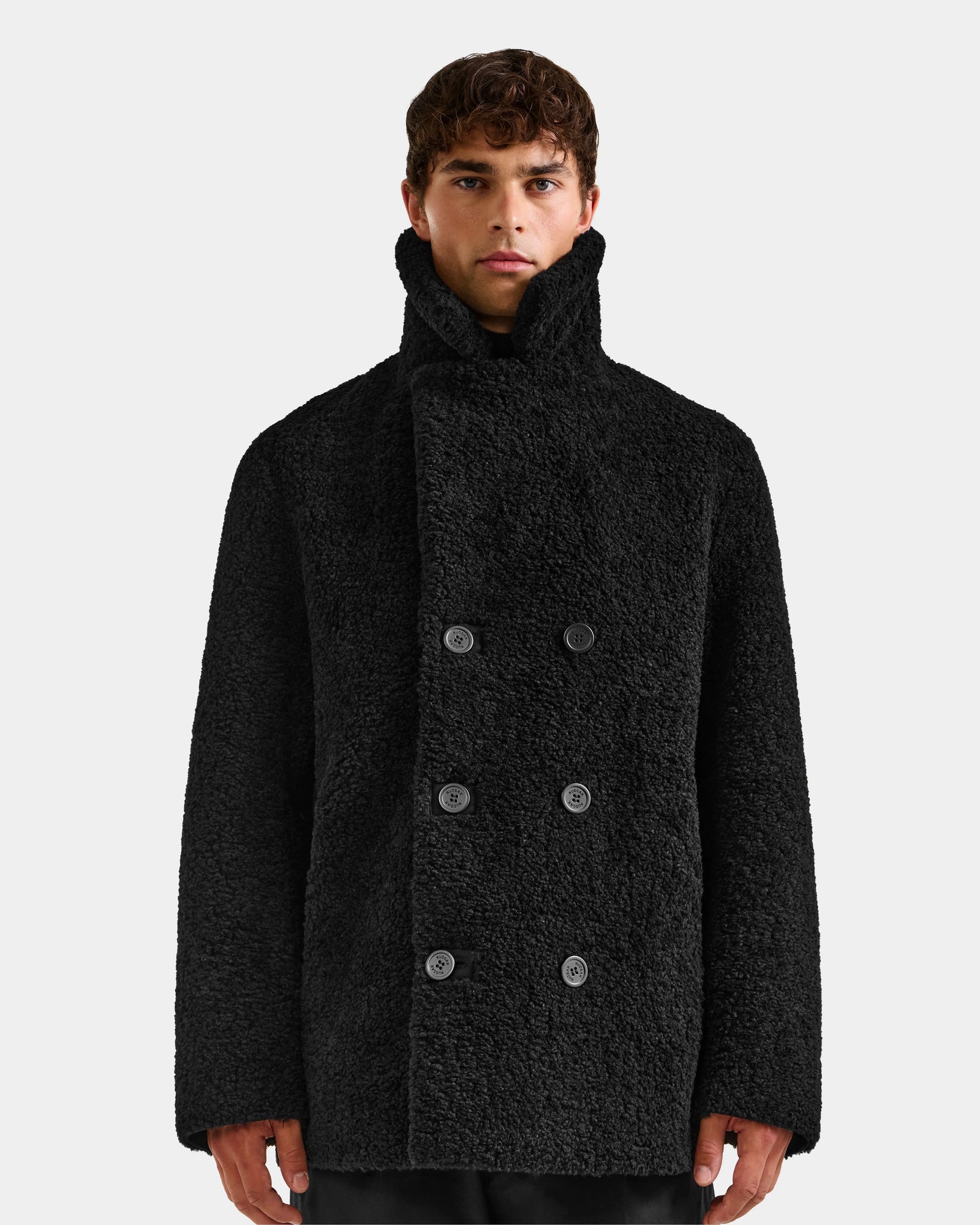 A person wearing the Rudsak ELTON - M125534 Black double-breasted overcoat with a high collar stands against a plain light background, looking straight at the camera.