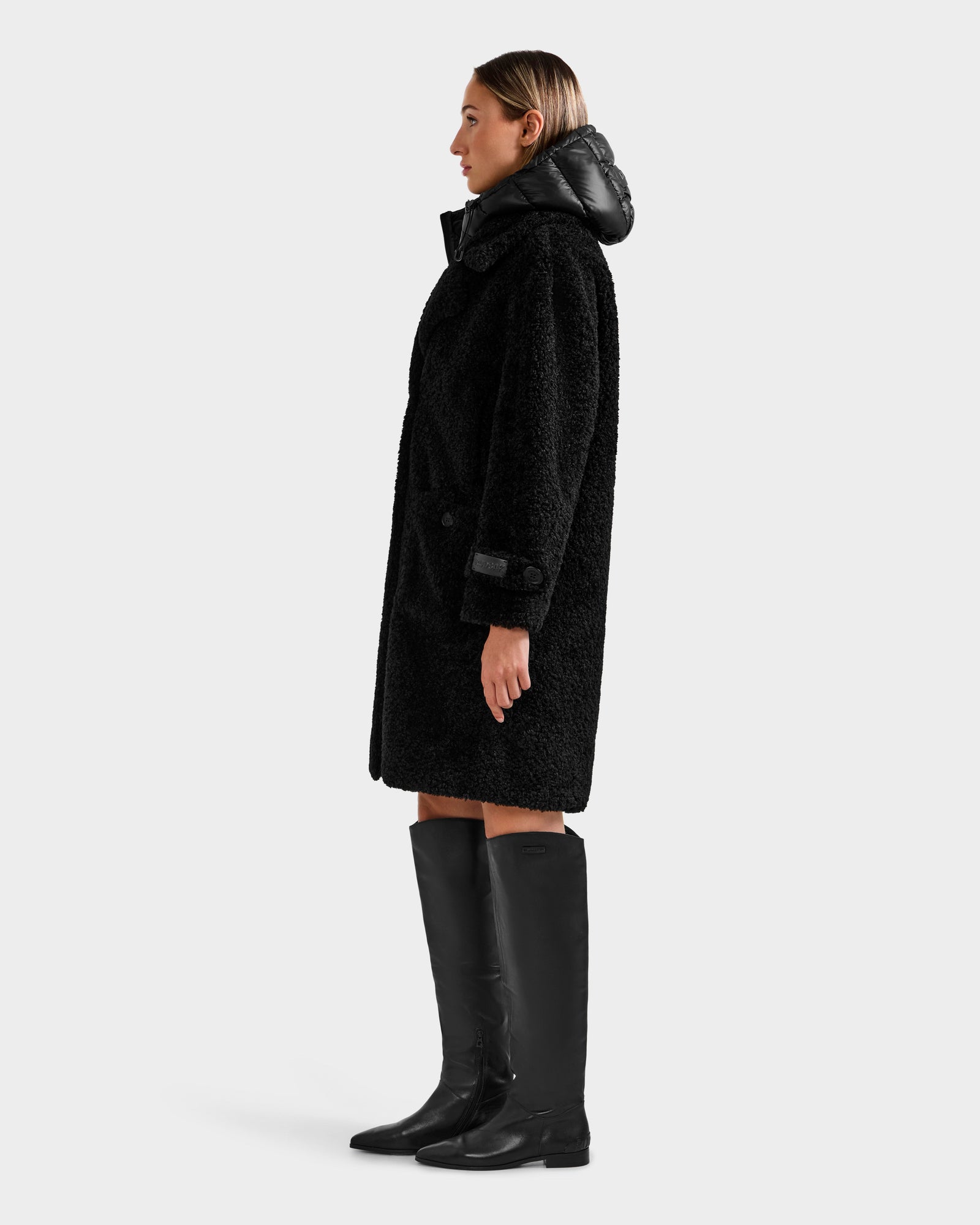 A person stands in profile wearing the Rudsak EMRYN - L125588 Black textured winter coat with a puffy collar and black knee-high boots against a plain light background.