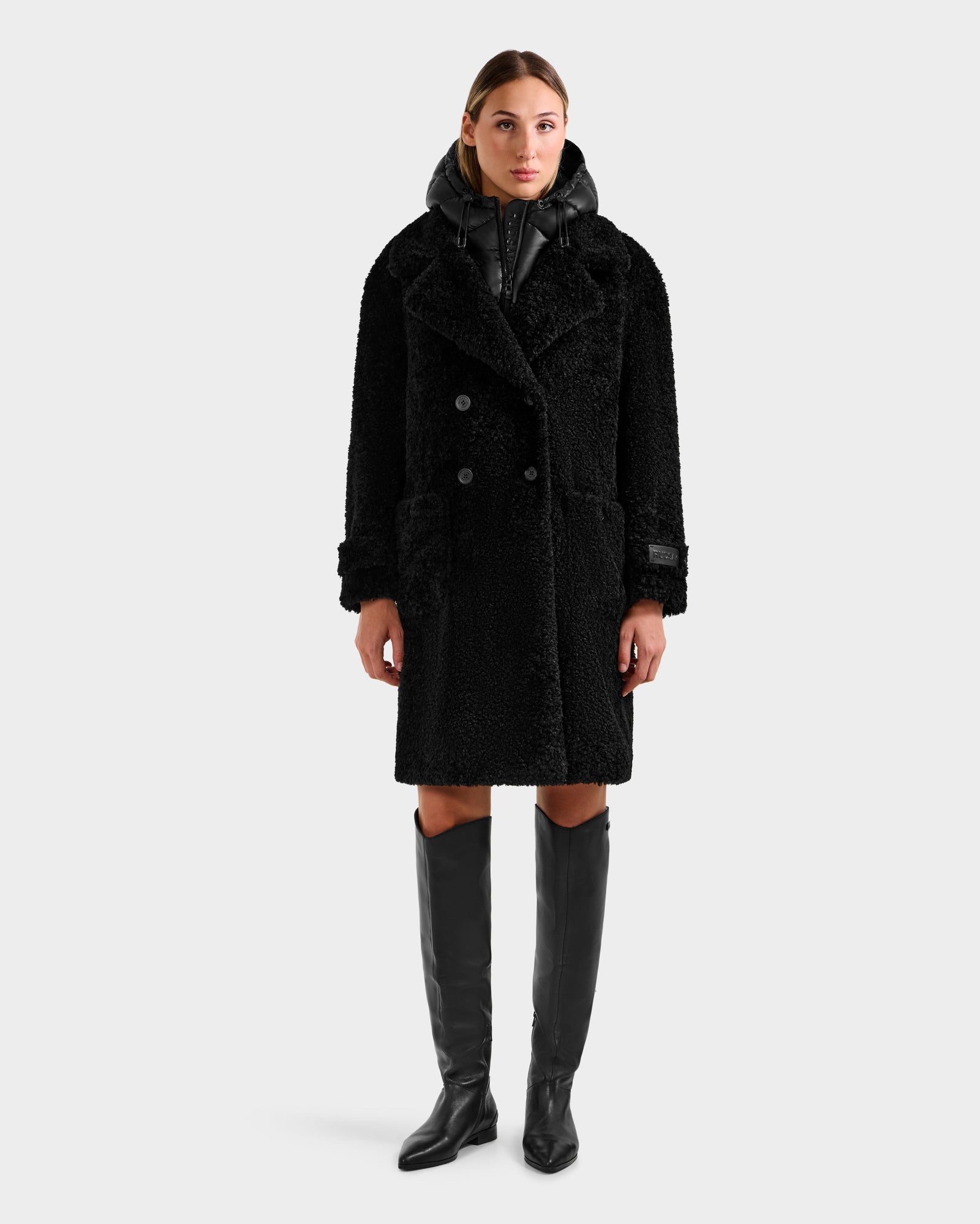A woman faces forward in a Rudsak EMRYN - L125588 Black textured winter coat with large lapels, layered over a black hoodie and styled with knee-high black boots against a plain light gray background.