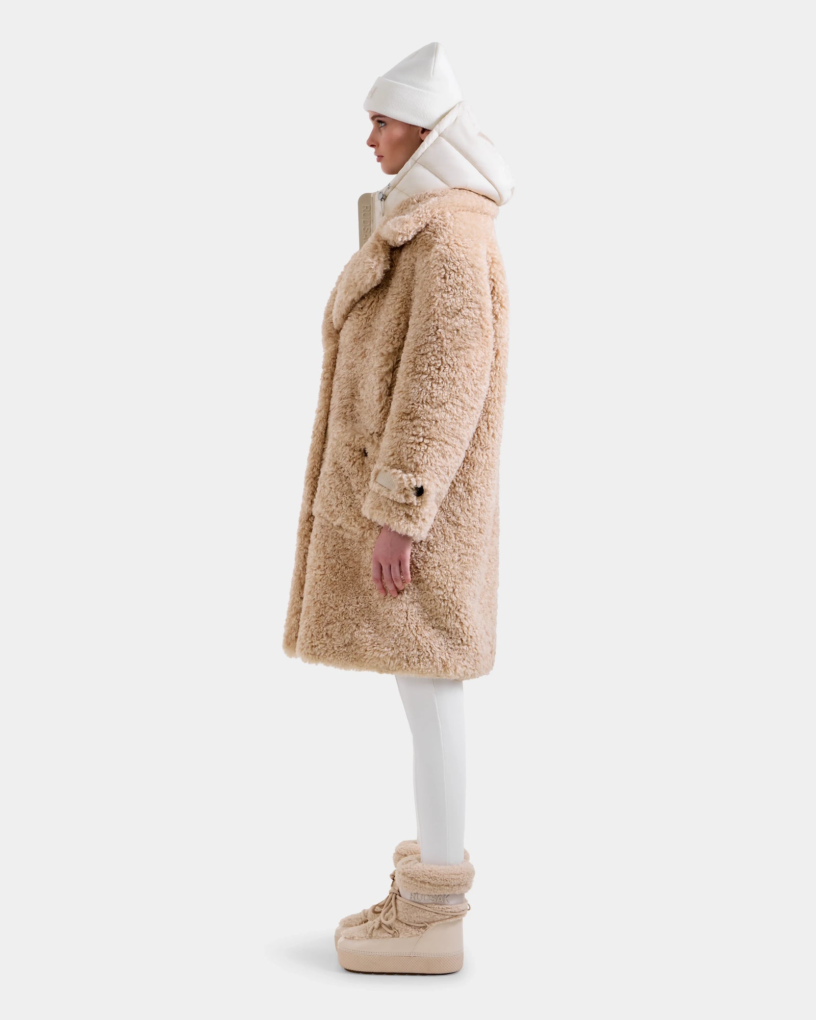 A person stands in profile against a plain light background, wearing the Rudsak EMRYN - L125588 Vanilla women's teddy jacket, paired with white leggings, beige fur boots, a white hooded top, and a white beanie.
