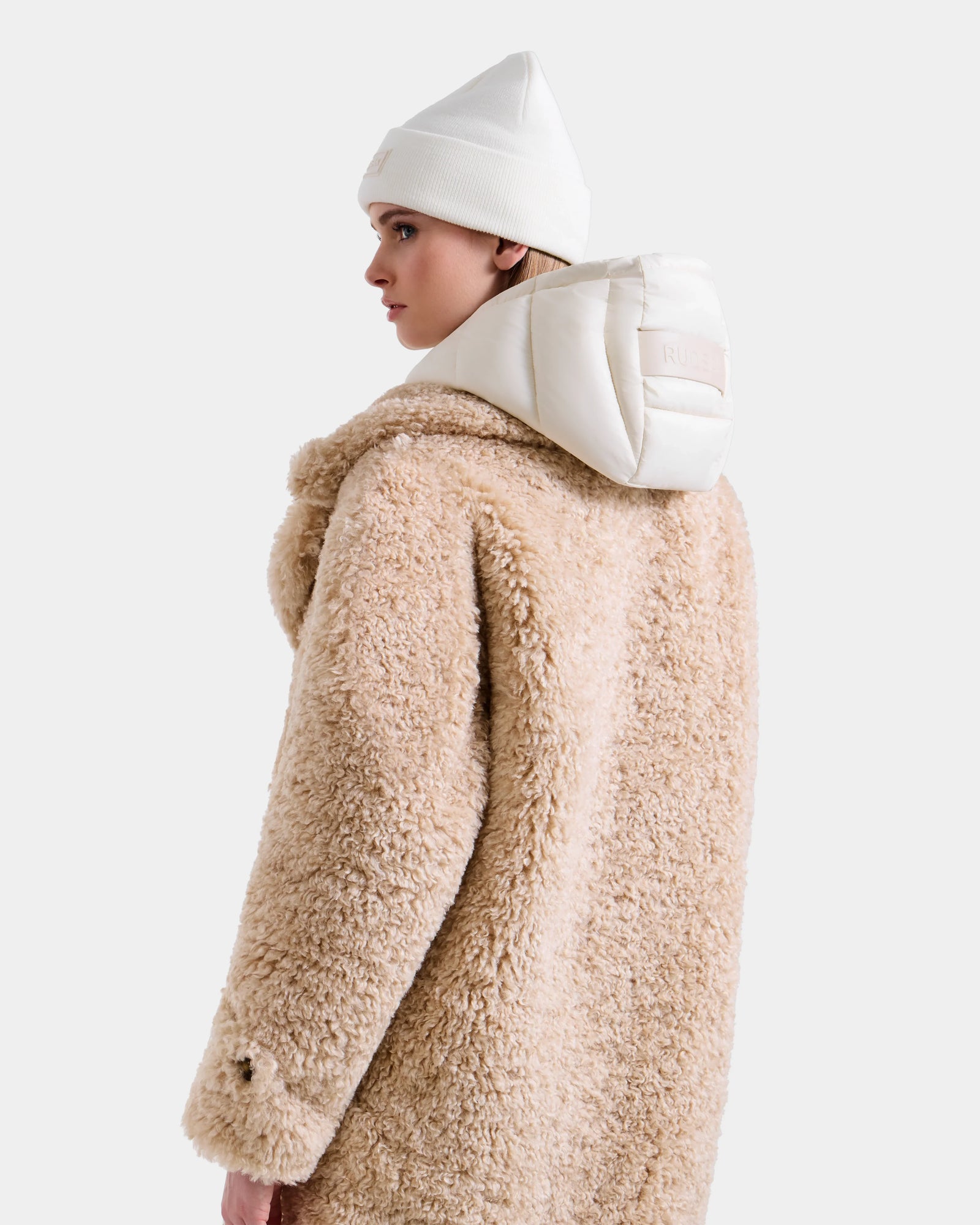 A person is seen sideways against a plain light background wearing the Rudsak EMRYN - L125588 Vanilla women’s teddy jacket, styled with a white padded hood and a knit beanie.