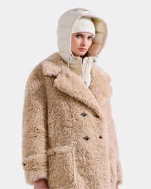 A person wears the Rudsak EMRYN - L125588 Vanilla teddy jacket, featuring large pockets and black buttons, paired with a cream puffer hood and beanie, standing against a white background.