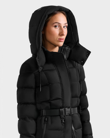 Wearing the Rudsak FRIDA M-L - L125608 Black, a person stands slightly to the side in front of a plain light background, showing off the quilted down coat’s belted waist and detachable hood.