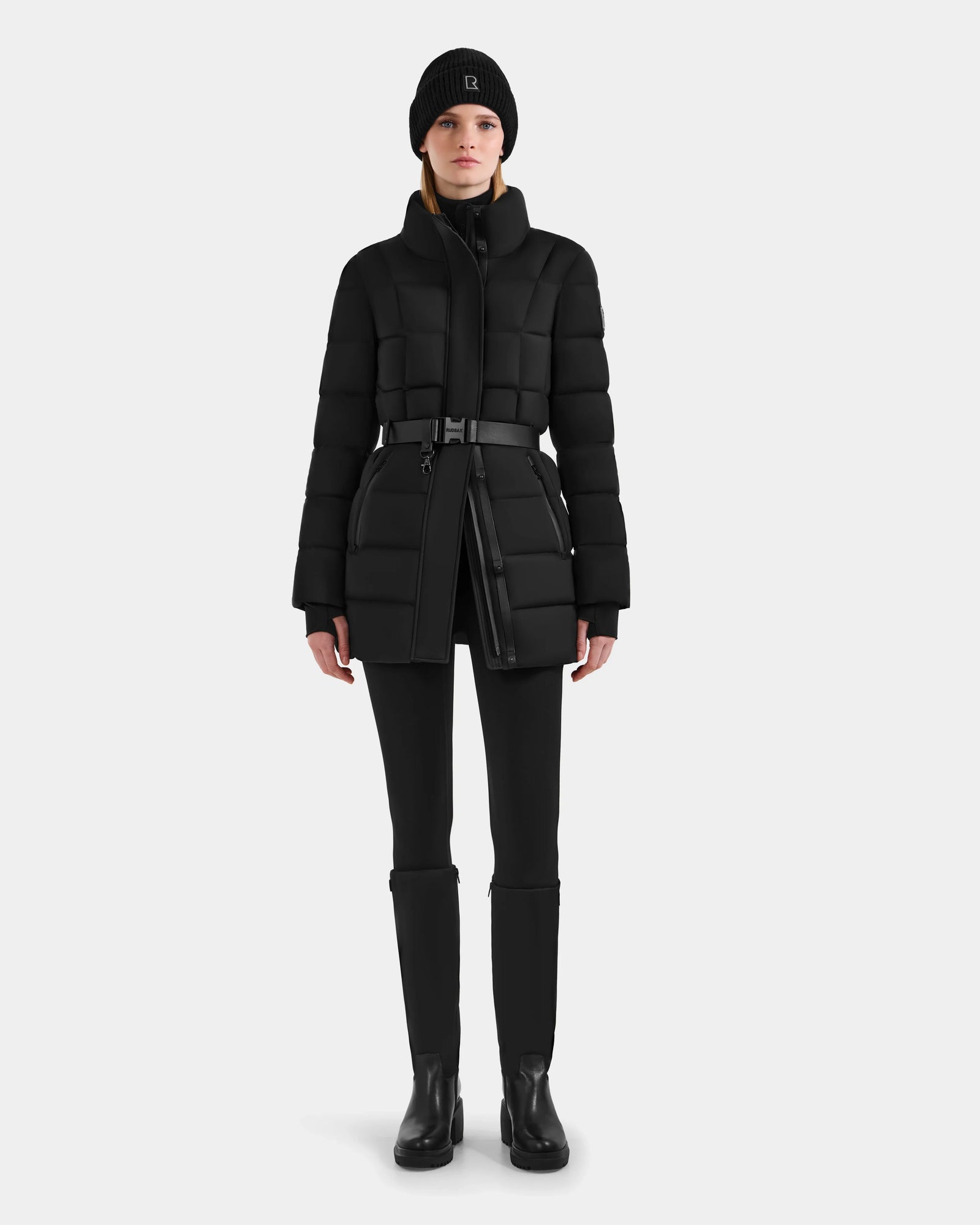 Wearing the Rudsak FRIDA M-M - L125607 Black women's quilted down puffer with a detachable hood, paired with black pants, boots, and a beanie, she stands facing forward against a white background—a classic winter essential.