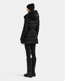 Dressed in the Rudsak FRIDA M-M - L125607 Black quilted down puffer with detachable hood, a black knit hat, black leggings, and knee-high boots, a woman stands facing away against a plain light background.