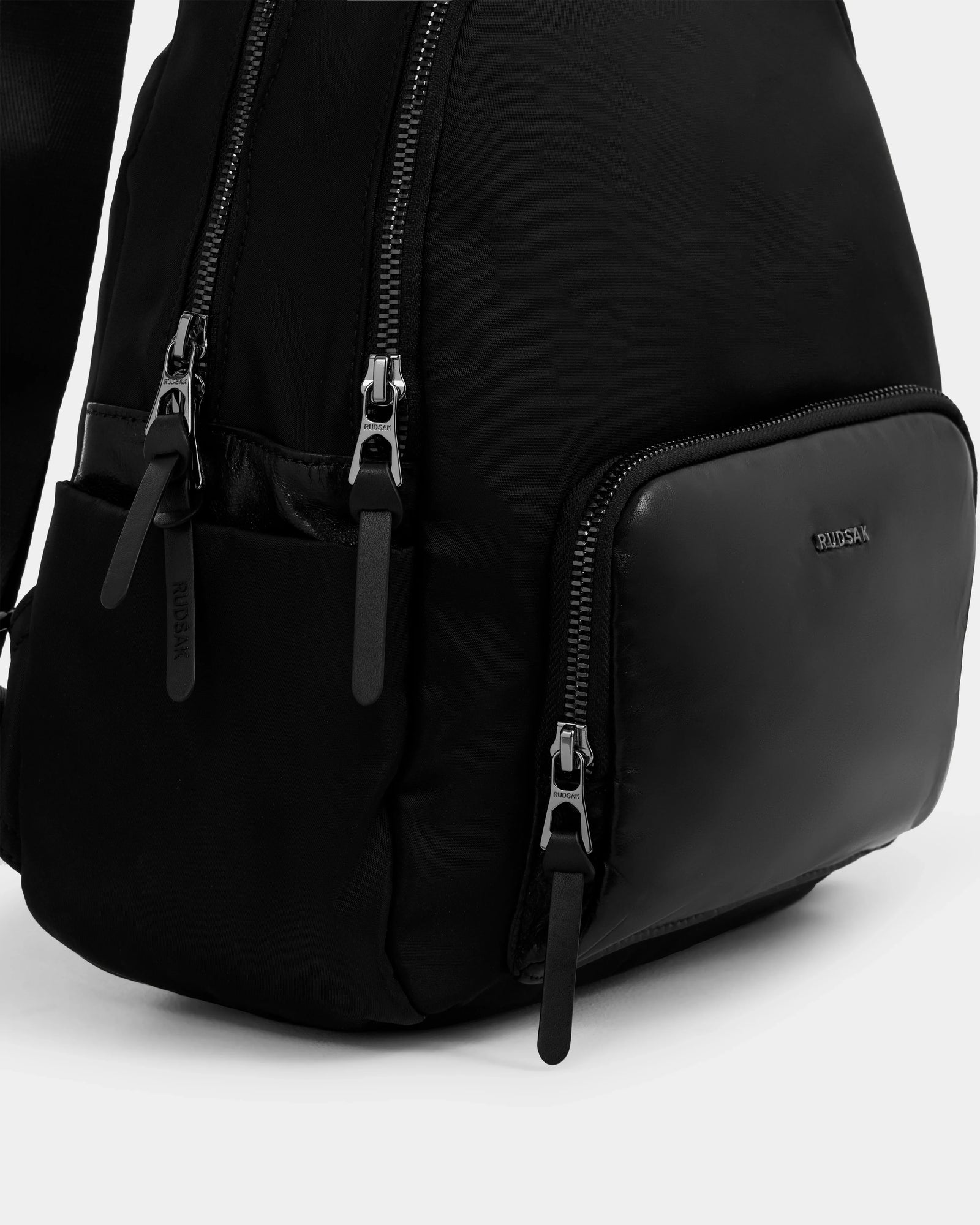 Close-up of the GRACE - L325013 Black women’s mini backpack by Rudsak, featuring multiple zippered compartments, a smooth front pocket, and a branded metal plate—a city-ready essential crafted from lightweight technical fabric.