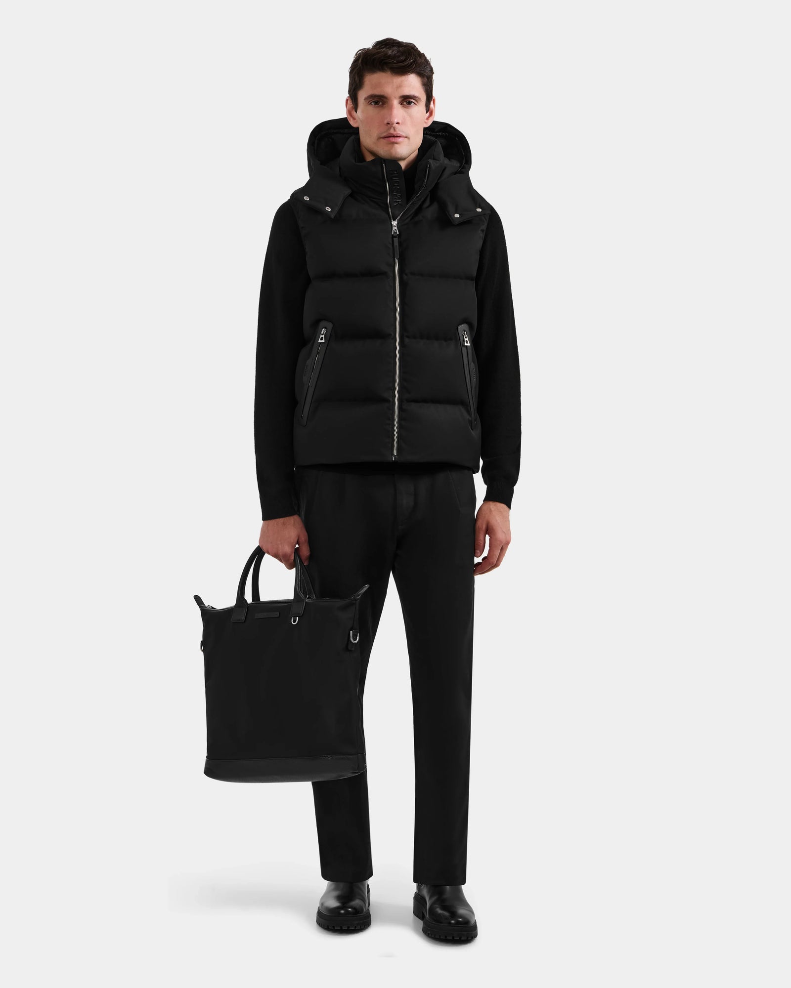 A man stands facing forward wearing the Rudsak HARLOW - M125528 Black men’s puffer vest with a removable hood over black clothes and holding a black tote bag in his left hand against a plain white background.