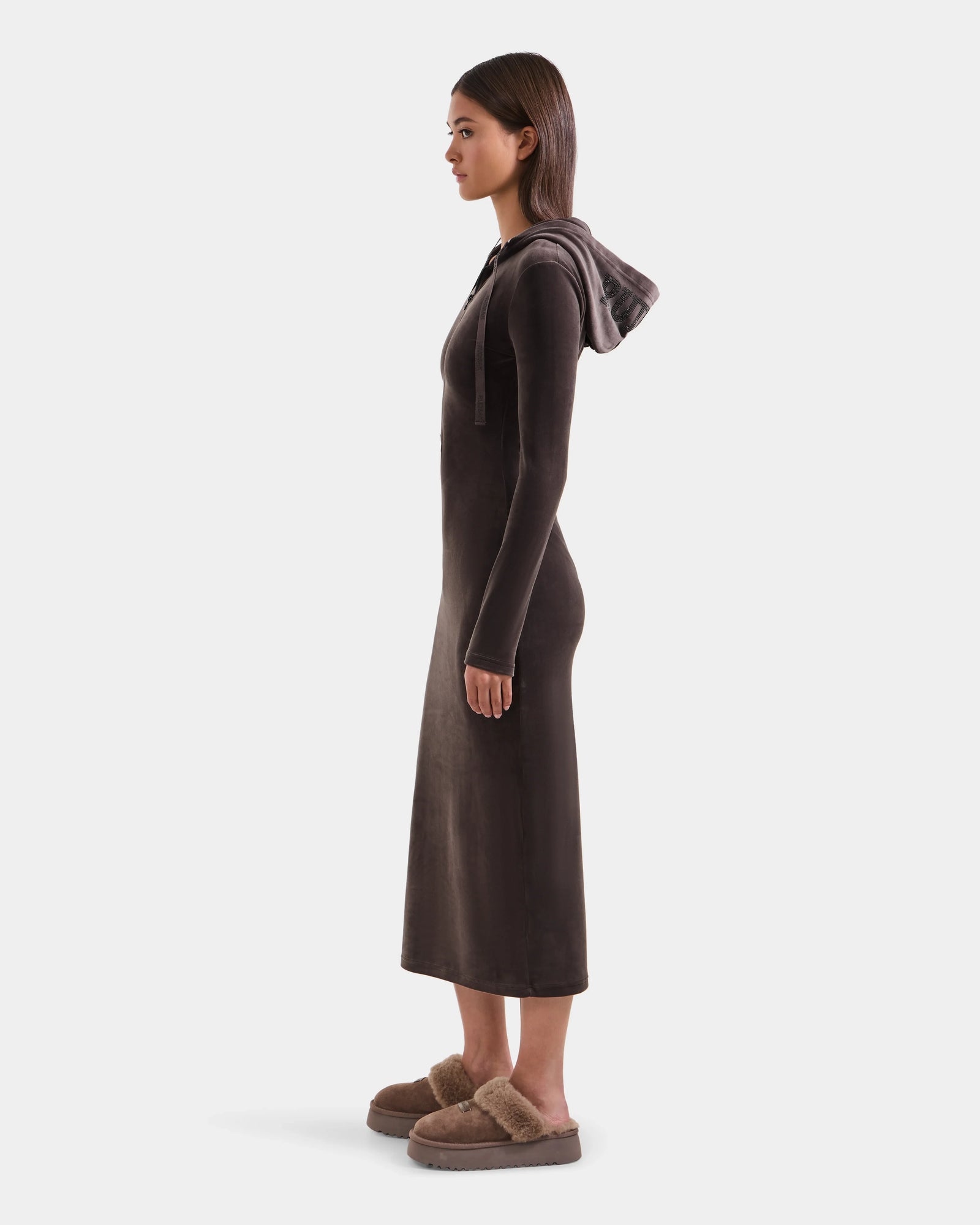 A woman in profile wears the Rudsak ISABELLA - L525502 Espresso, a long, dark brown velour hooded midi dress, paired with brown fuzzy slippers, set against a plain white background.