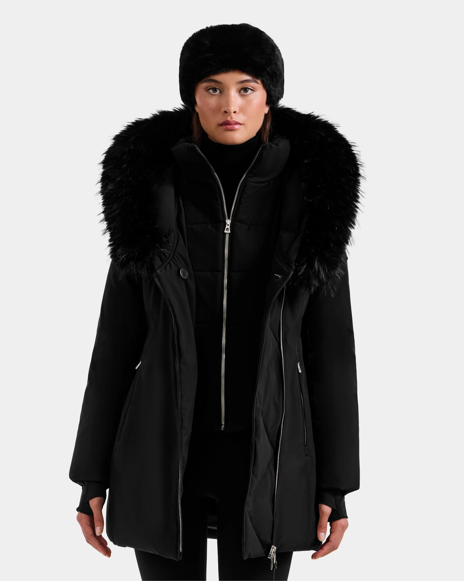 A woman with straight brown hair and a neutral expression stands against a plain light background, wearing the Rudsak ISELLE - L125547 Black down parka with a large faux fur-trimmed hood and a black fur hat.