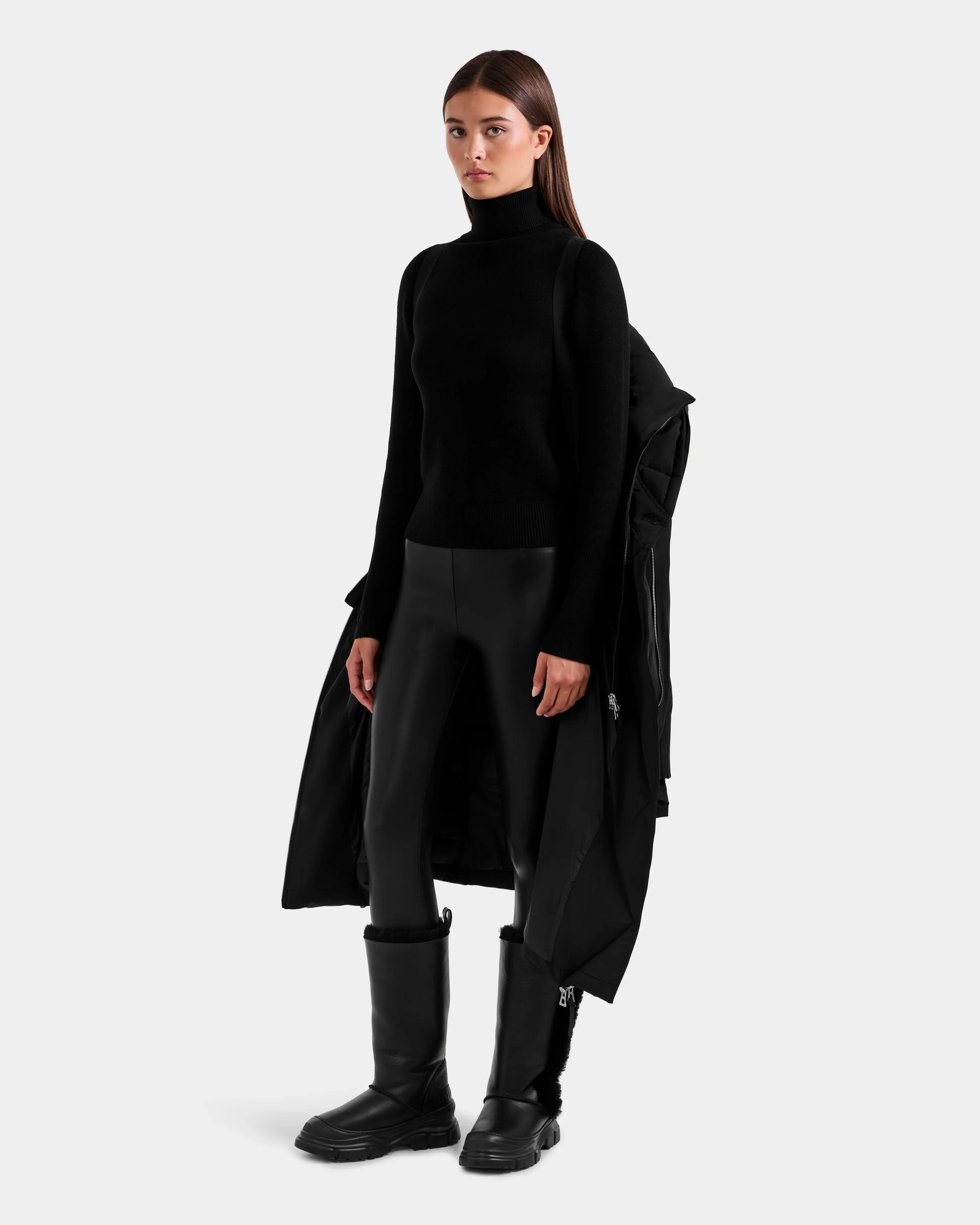 Dressed in the Rudsak JANIS - L125558 Black turtleneck and faux leather leggings, a person stands against a light gray background with black boots and a winter coat draped over one arm.