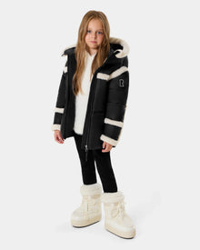 A young girl with long light brown hair stands facing forward, wearing the Rudsak JULIANA K - K125550 Black puffer jacket, black leggings, a white sweater, and white furry boots against a plain light background.