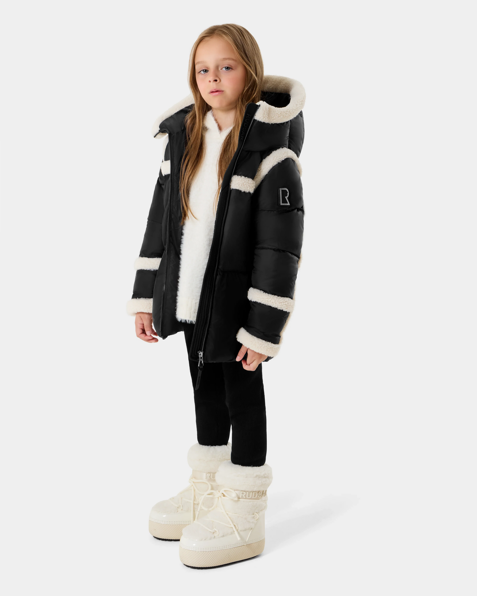 A young girl with long blonde hair wears the Rudsak JULIANA K - K125550 Black puffer jacket with white faux fur trim, paired with black leggings and white winter boots, posing against a plain light background.