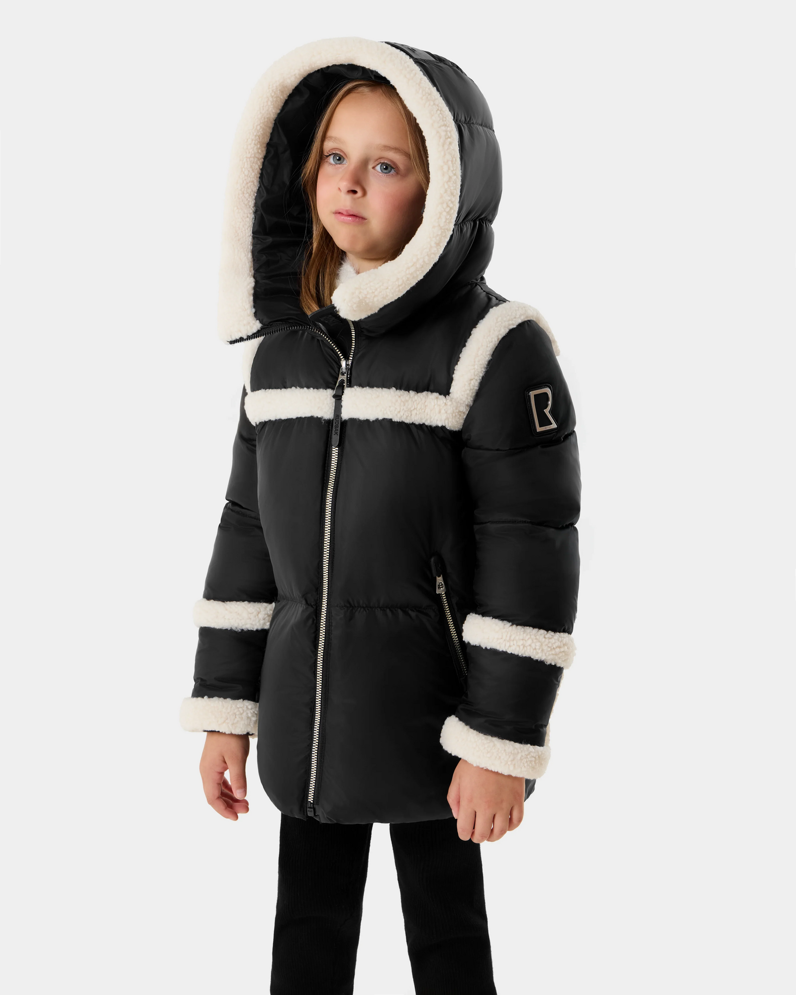 A young girl wears the Rudsak JULIANA K - K125550 Black winter coat with white faux fur trim, standing against a light gray background and gazing slightly upward and to the side.